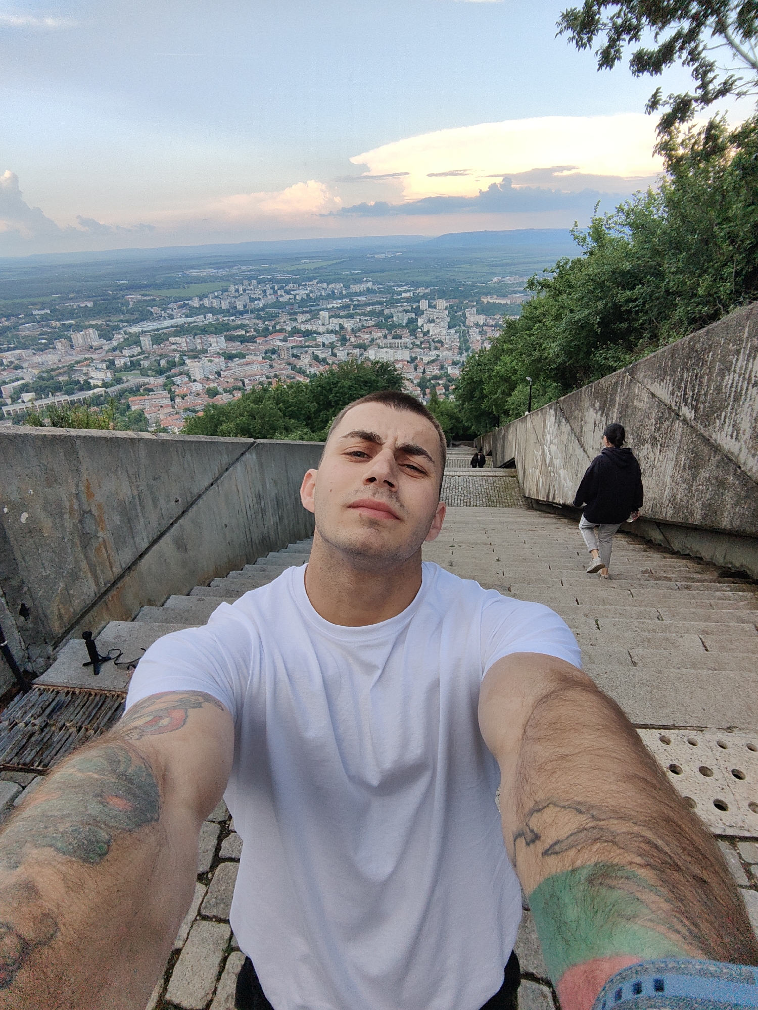A person with short brown hair and tattoos on both arms is taking a selfie on a stone staircase with a concrete wall to the left and right. They are wearing a white t-shirt. In the background, a city with numerous buildings stretches into the distance under a cloudy sky. There are people further down the stairs. To the right of the stairs is a green hillside. The photograph appears to be taken from near the Monument to 1300 Years of Bulgaria, overlooking the city of Shumen, Bulgaria.