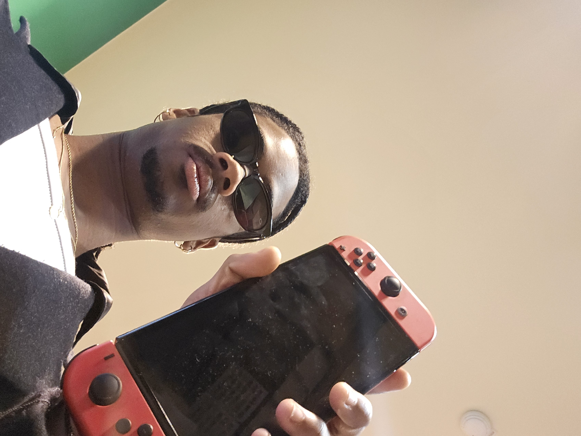 A person wearing sunglasses holds a Nintendo Switch console. The background features a green wall and a beige ceiling. There are no indications this image was taken in Paris.