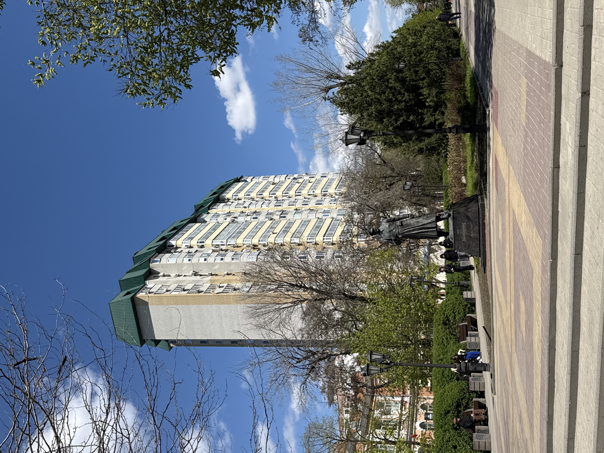 The image shows a tall, multi-story building in Rostov-on-Don, Russia, identifiable by its distinctive green roof and light-colored facade with numerous windows and air conditioning units. The building is set against a bright blue sky with scattered clouds. In the foreground, there are bare trees with branches reaching into the frame. To the right, a section of a pedestrian walkway made of paving stones and concrete steps is visible. A statue stands in the middle ground, along with lampposts and greenery, including bushes and trees. Benches can be seen near the walkway, and people are sitting on them. The scene appears to be taken in a park or public space.