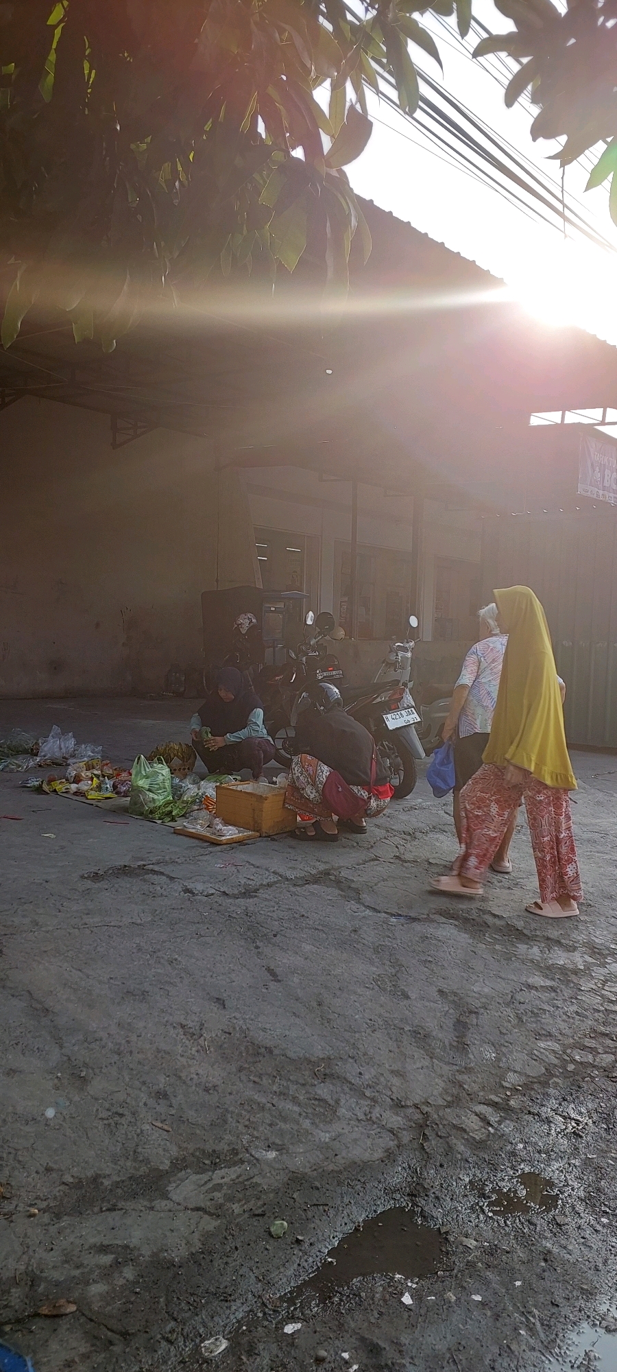 The image depicts an outdoor scene in what appears to be a market or a roadside vending area in Semarang, Indonesia, indicated by the style of the building and the motorcycles commonly used for transport. The photograph is taken at an angle, with the sun creating a lens flare effect in the upper left quadrant, obscuring some details.
In the foreground, the ground is a rough, grey concrete surface with visible cracks and some puddles of water, suggesting recent rain or a damp environment. Scattered on the ground to the left is an assortment of goods, including plastic bags filled with produce, loose fruits and vegetables, and a wooden crate.
Several individuals are present in the middle ground. Two people are crouched down near the scattered goods, appearing to be vendors or shoppers interacting with the merchandise. One person is wearing a dark hijab and a light-colored top, while the other is wearing a patterned skirt and a dark top. A third person, dressed in a yellow hijab and patterned pants, is standing and walking away from the viewer towards the right side of the image. Another individual, seen from the back, is wearing a patterned shirt and is also facing away.
Behind the people and goods, several motorcycles are parked. The background features a low-slung building with an overhang, typical of small businesses or market stalls. The overall lighting suggests it is either early morning or late afternoon, with the sun's glare contributing to the atmospheric haze.