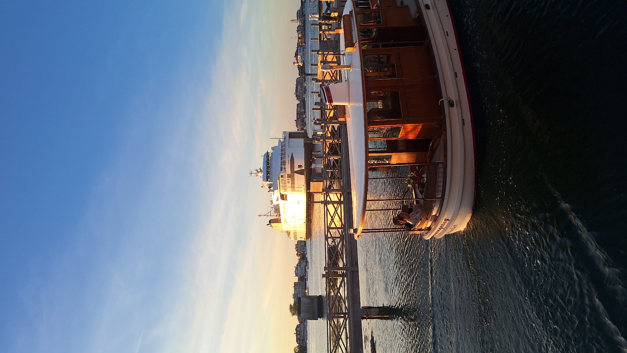 The image displays a serene harbor scene during the golden hour, likely sunset, in Nantucket, United States of America. A classic wooden boat, identified by the name "Annaburg N.Y." on its stern, dominates the right side of the frame. This vessel features a white hull with a red stripe and a rich wooden superstructure with windows. Two individuals are visible relaxing on its stern deck, seemingly enjoying the peaceful setting.
In the mid-ground, a large white ferry or cruise ship is docked or anchored further down the waterway, along with numerous other smaller boats and residential buildings lining the distant shore. A wooden pier or dock structure, composed of pylons and horizontal beams, extends from the foreground towards these distant vessels. The sky is a clear, vibrant blue, transitioning into warm orange and yellow tones near the horizon due to the low sun. The sunlight creates a shimmering reflection on the calm water, which shows gentle ripples and a slight wake behind the "Annaburg," indicating slow movement or recent activity. The overall ambiance is tranquil and picturesque.