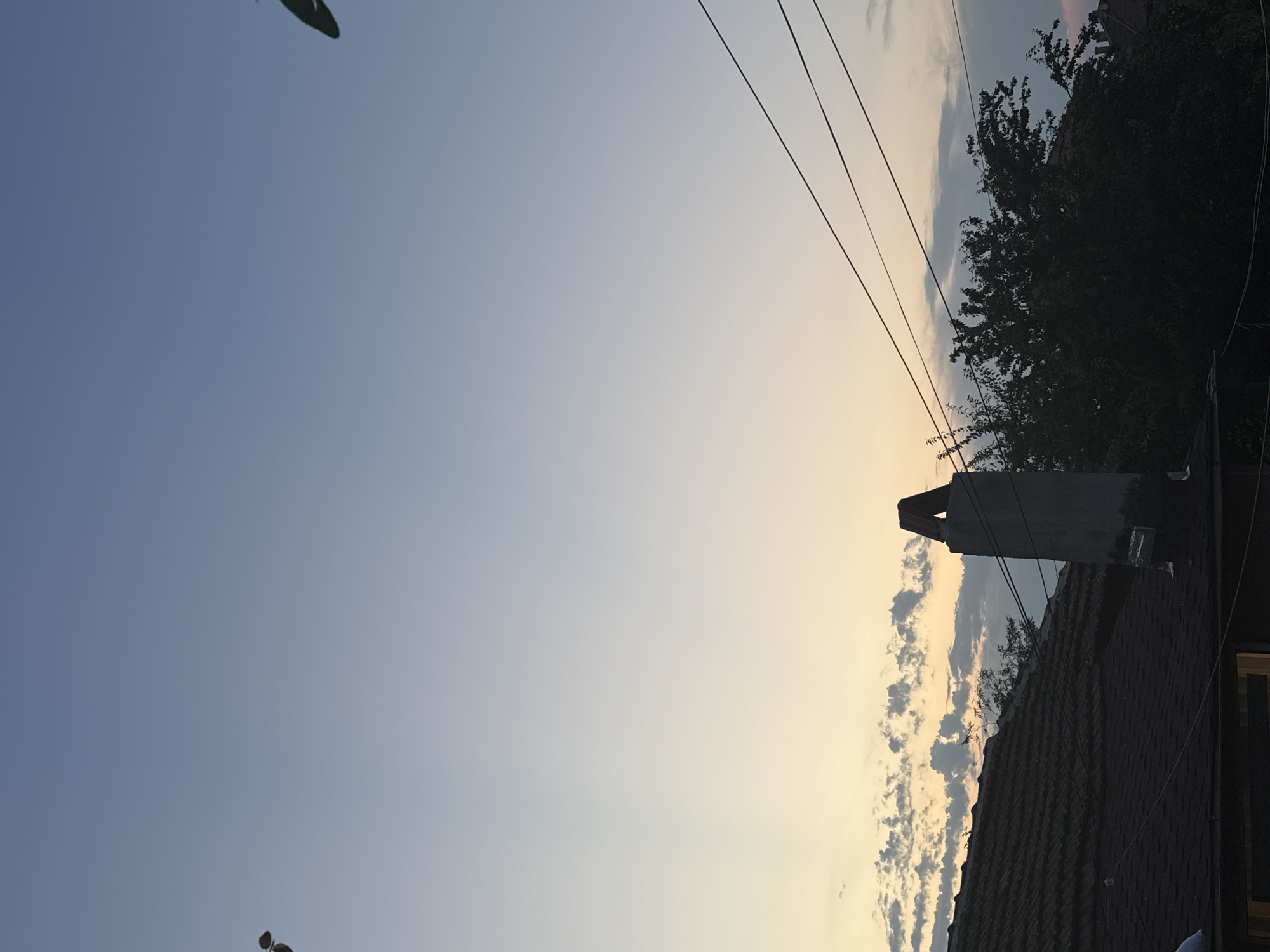The image depicts a rooftop view at sunset in Sredets, Bulgaria. The sky transitions from a pale blue at the top to a soft yellow and white near the horizon, with scattered, light grey clouds. In the foreground, a dark tiled roof occupies the lower left portion of the frame. To the right of the roof, a tall, grey chimney with a triangular cap stands prominently. Behind the chimney and to the right, dense, dark green foliage of trees partially obscures more rooftops and structures in the distance. Several black electrical wires crisscross the sky horizontally. The scene is devoid of any people or animals.