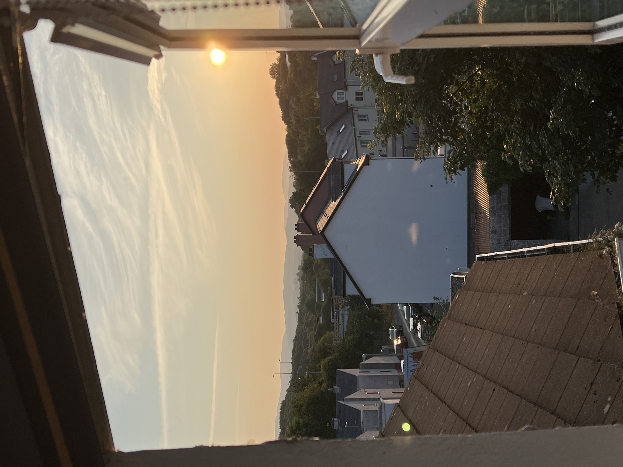 This is a photograph taken from an elevated vantage point, looking out over a residential area at sunset. The foreground is dominated by the edge of a window frame and a portion of a roof. The sky is a warm orange and yellow gradient, with wispy clouds stretching across it. In the middle ground, several houses with red-tiled roofs are visible, along with trees and a street with parked cars. The background consists of rolling hills and distant mountains, silhouetted against the setting sun. The sun itself is a bright orb, positioned towards the right side of the frame, casting a warm glow over the scene. The overall impression is one of a peaceful evening in a town.