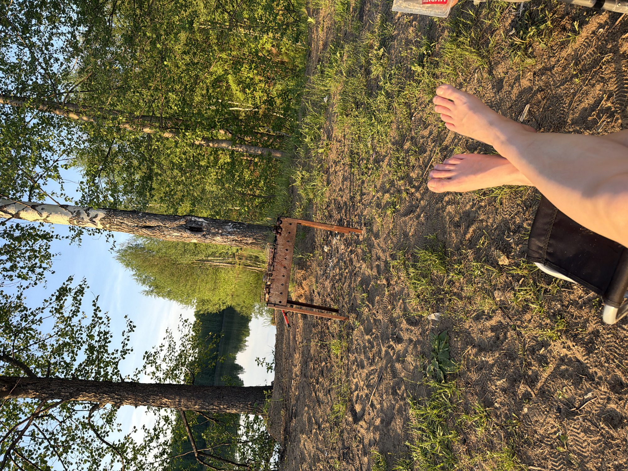 The image shows a person's bare feet resting on sandy ground next to a portable grill, indicating an outdoor recreational setting. The location appears to be near a lake or river, as evidenced by the visible water and surrounding trees, suggesting a natural or park area within the city of Unknown, Unknown.
