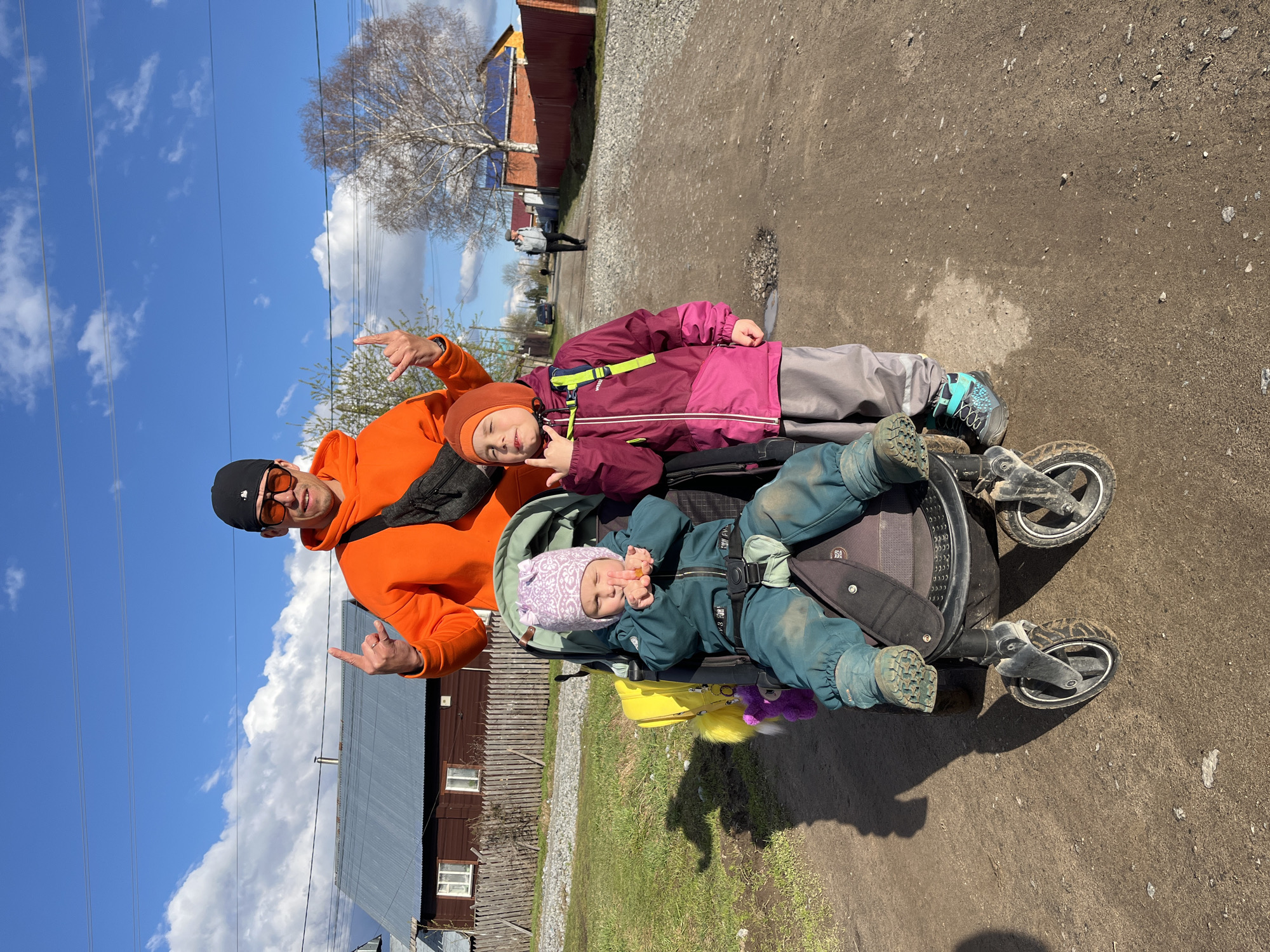 An adult male and two children pose playfully outdoors on what appears to be a sunny day in Chaikovsky, Russia. The image is taken on a dirt and gravel path, with residential buildings and natural elements in the background.
The adult male, on the left, wears a bright orange hoodie, a black baseball cap, and orange-tinted sunglasses. He has a black fanny pack slung across his chest. He faces the camera, smiling, and makes a "rock on" hand gesture with both hands.
To his right stands an older child, wearing a burgundy and grey winter jacket, grey trousers, and an orange knit hat. This child also smiles at the camera and mimics the "rock on" gesture with their right hand. Their shoes are light teal and black.
A younger child sits in a dark-colored stroller on the right side of the frame. This child wears a dark teal full-body suit, a light purple patterned hat, and appears to be holding a pacifier or small object in their mouth while also raising a hand in a similar gesture. Their suit and boots show signs of dirt. A yellow bag and a purple plush toy are attached to the stroller.
The background features a clear blue sky with scattered white clouds and visible power lines. To the left, a bare deciduous tree suggests early spring or late autumn. To the right, a wooden fence and a rustic wooden house with a corrugated metal roof are visible. Further down the path, another person is seen walking in the distance. The bright sunlight casts clear shadows, indicating midday. The overall mood is cheerful and relaxed. No discernible text is visible on clothing or objects.