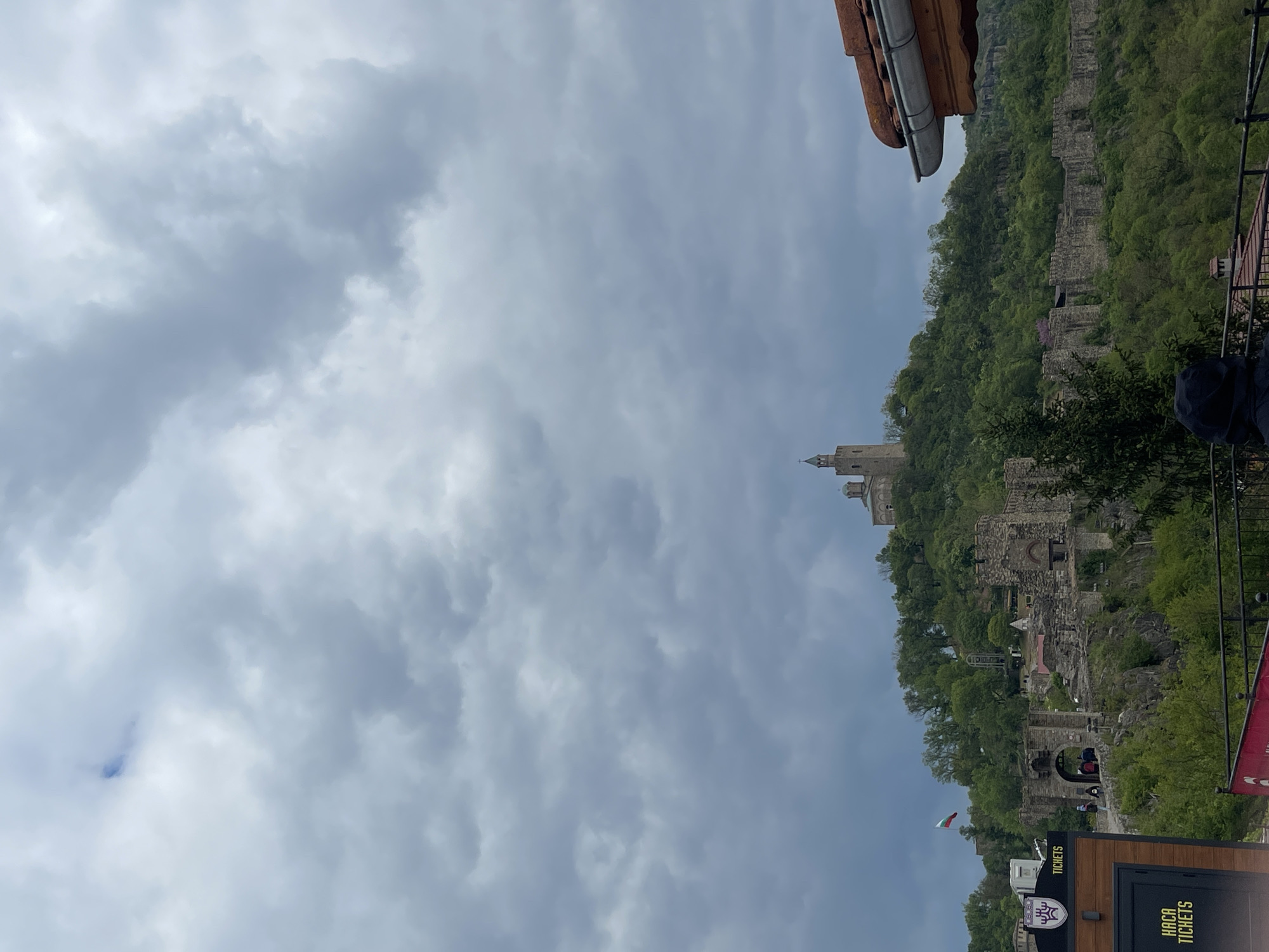 The image captures a view of Tsarevets Fortress in Veliko Tarnovo, Bulgaria, under a cloudy sky. The ancient fortress, situated on a hill covered in trees, includes visible stone walls, ruins, and a tower. In the foreground, there is some signage for "Tickets".