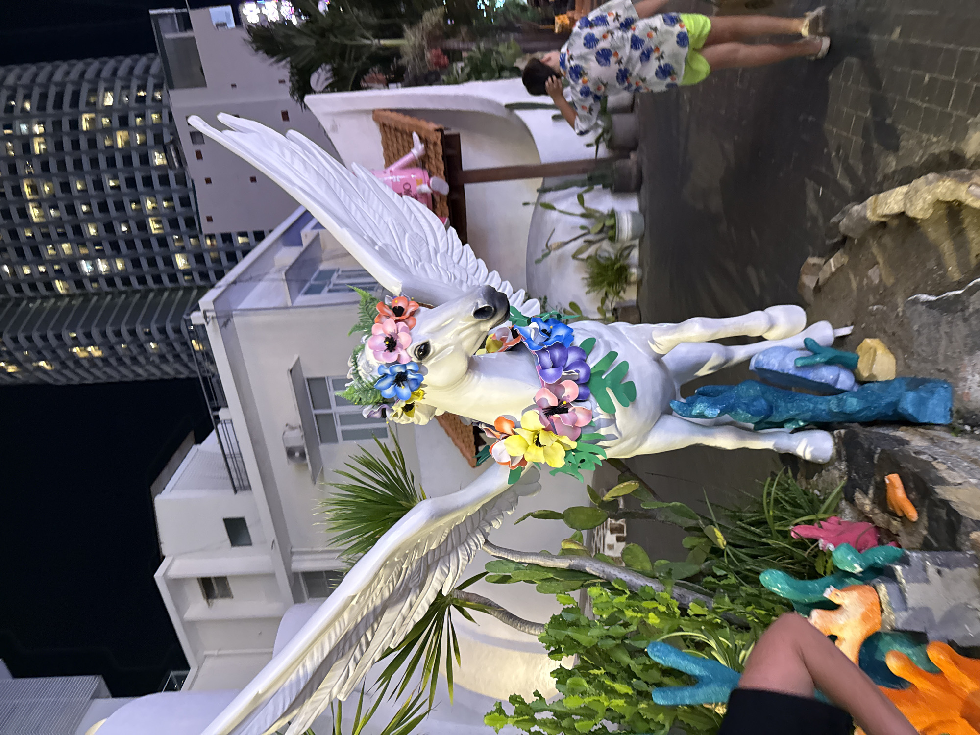 An outdoor nighttime scene in Nha Trang, Vietnam, features a white statue of a winged horse adorned with a colorful flower garland. The statue stands on a paved area amidst various potted plants, including cacti and palm trees, and some brightly colored, abstract sculptures. In the background, a modern high-rise building with many illuminated windows is visible, along with a white building structure. To the right of the statue, a person wearing a patterned shirt and bright green shorts is walking away from the camera, adjusting their hair. Another person's arm and shoulder, wearing a black shirt, are visible on the far left, interacting with the colorful sculptures. The lighting suggests it is evening or night.