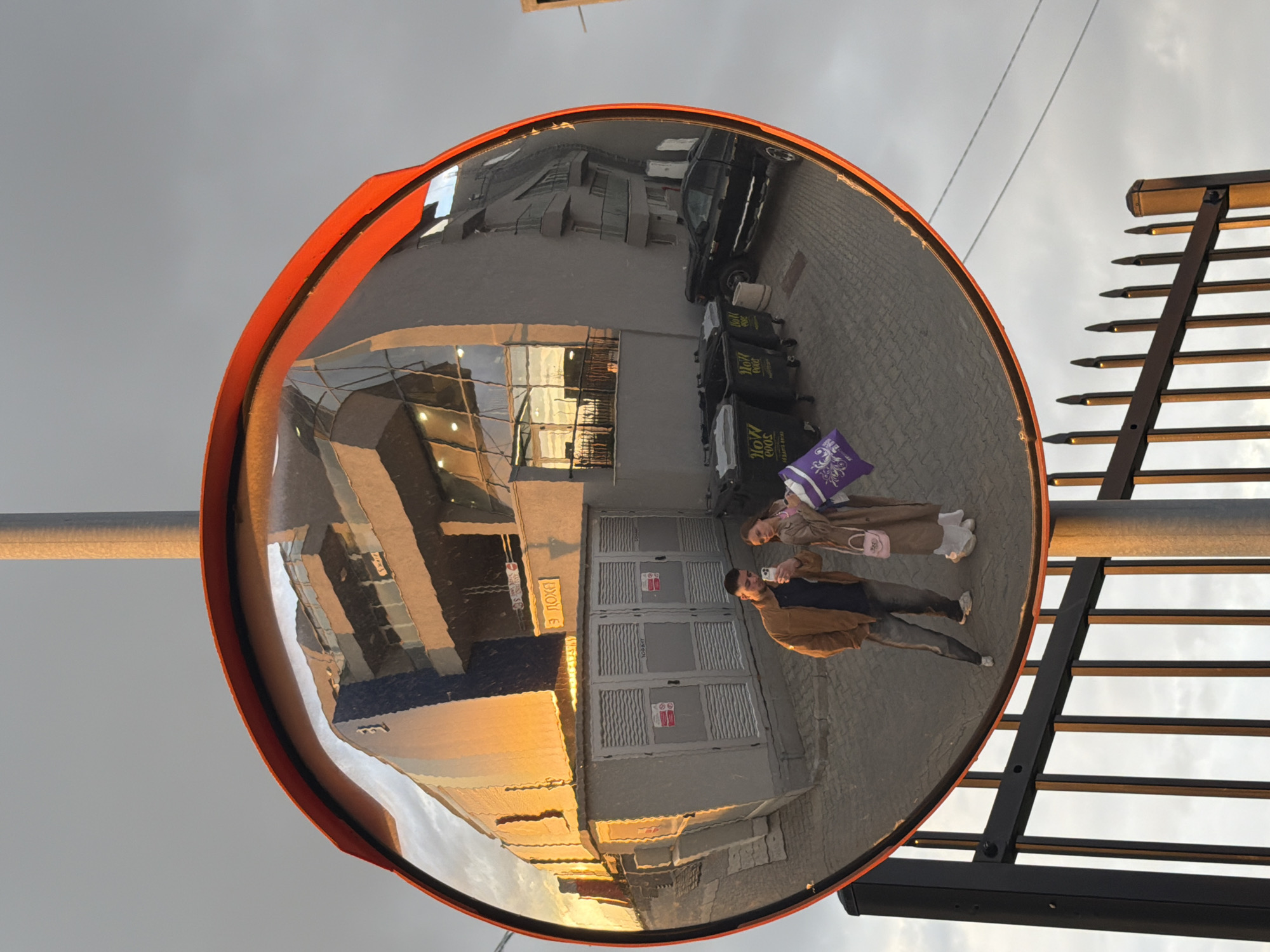 The image displays an urban scene in Sofia, Bulgaria, captured through the distorting reflection of a large, circular convex mirror with a bright orange rim. The mirror is affixed to a metal pole on the left, while a black metal fence or railing is visible on the right, silhouetted against a largely overcast sky.
Within the mirror's reflection, two people are central subjects. A man, dressed in a brown jacket and dark trousers, holds a smartphone, potentially taking a picture or video. Beside him, a woman wearing a light brown coat and white sneakers carries a small pink handbag and a purple tote bag. They stand on paved ground, possibly cobblestones or pavers.
The reflection encompasses a broader view of the surroundings: buildings with numerous windows, some reflecting warm, golden light indicative of either sunrise or sunset. A dark-colored car is parked on the street, and several black trash bins are visible, marked with Cyrillic text including "СОФИЯ" (Sofia) and "ЕКО" (Eco), alongside "NEW". A sign on one of the reflected buildings shows the text "1937" and "ЕДНО ПОХОД", likely an address or street name. The lighting suggests late afternoon or early morning, with the golden hues on the buildings creating a notable detail.