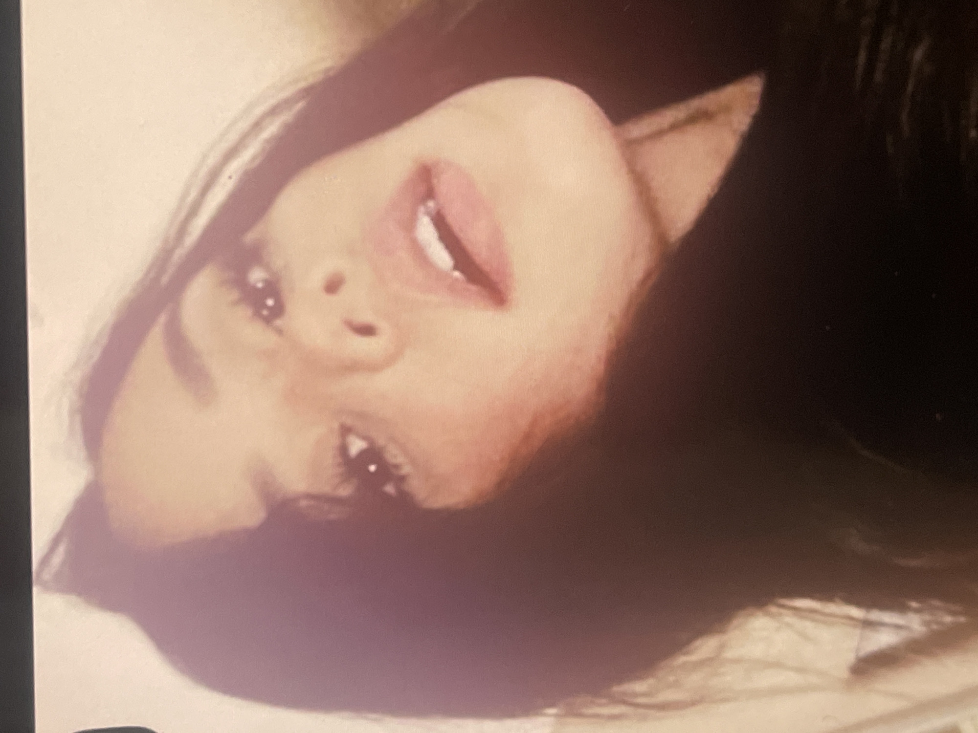 This image is a close-up portrait of a young woman, captured with her head tilted sharply to the right. She has long, dark hair, which is visible on the right side of the frame and appears to be flowing around her. Her face, characterized by a fair complexion, is the central focus. Her lips are slightly parted, revealing the upper edge of her teeth, and are adorned with what appears to be a light pink or nude lipstick. Her dark eyes are open and look towards the viewer, reflecting a soft light, and are accentuated by prominent eyelashes. The lighting is soft and warm, casting gentle shadows that highlight her facial contours and create a serene, almost contemplative expression. The background is largely out of focus and consists of muted, light tones, suggesting an indoor setting without any specific details or objects. There is no visible text in the image. While the image itself does not contain specific geographical cues, the portrait is contextualized as being from Poltava, Ukraine. The soft and intimate lighting suggests a controlled environment, rather than a specific time of day.