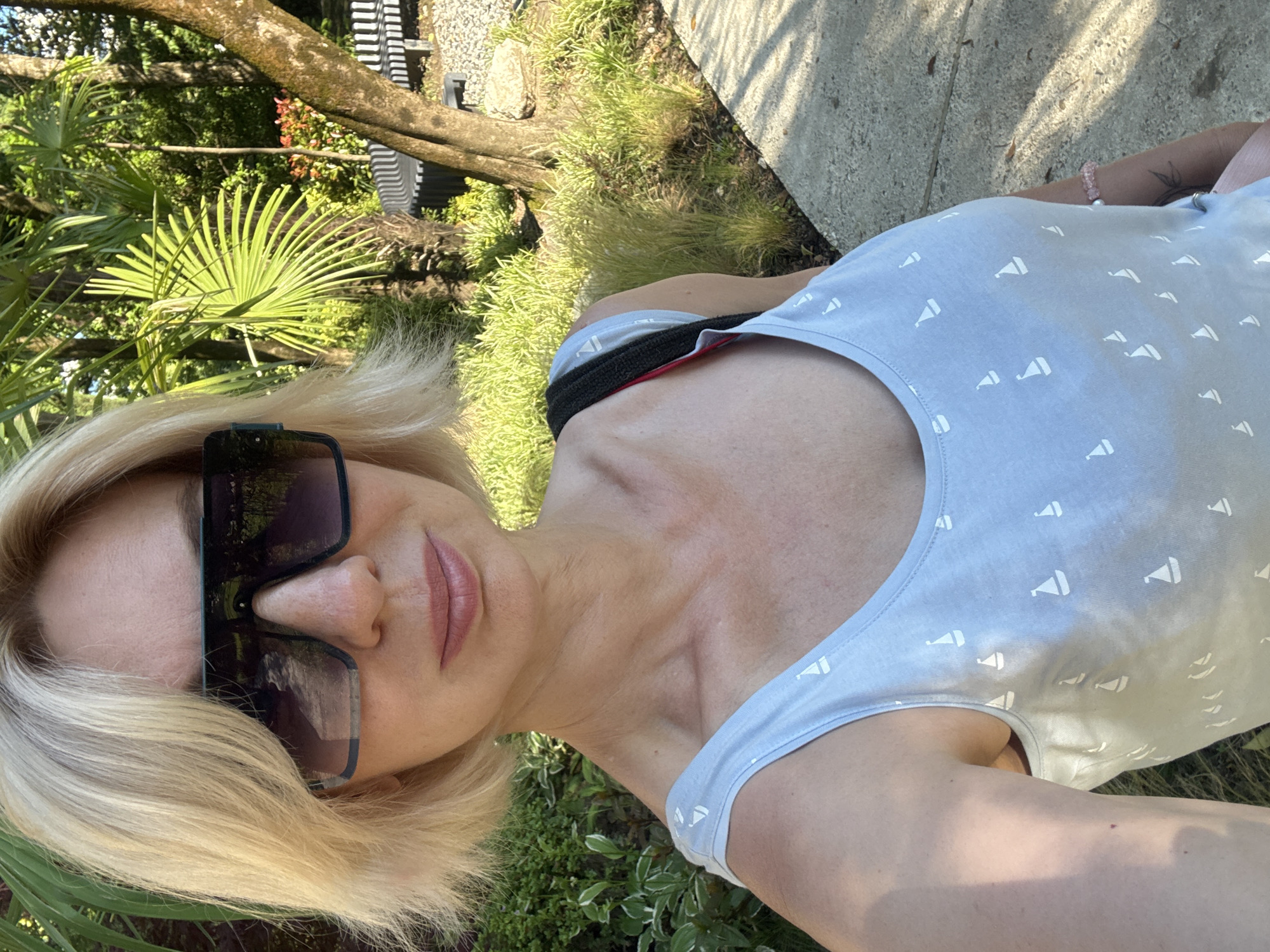 In Sochi, Russia, a fair-skinned woman with short blonde hair and sunglasses takes a selfie outdoors. She is wearing a light blue tank top with a pattern of small white sailboats. The background features lush green foliage, palm trees, and a paved pathway. A black and white striped bench is visible behind her. The overall scene suggests a warm, sunny day in a park or garden setting.