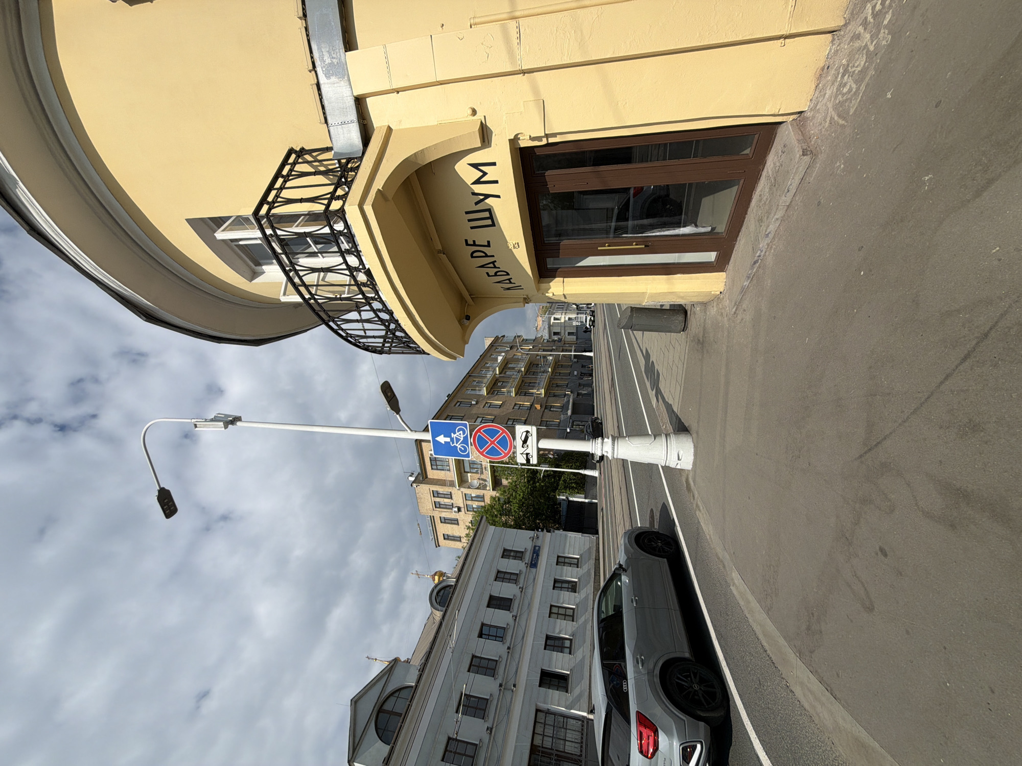 The image depicts a street scene in an urban location, possibly in Russia, given the presence of Cyrillic script on a building. The predominant element is a yellow building on the left side of the frame, featuring a curved façade with a black iron balcony. An awning with the inscription "КАБАРЕ ШУМ" (Cabaret Noise) covers an entrance. The street is paved, and a gray car is parked along the right curb. A white post with a blue traffic sign prohibiting bicycles and parking is positioned next to the car. Beyond the car and the traffic sign, a view of other buildings can be seen in the distance. The sky is overcast with light gray clouds. The image is oriented sideways, giving a rotated perspective of the street.