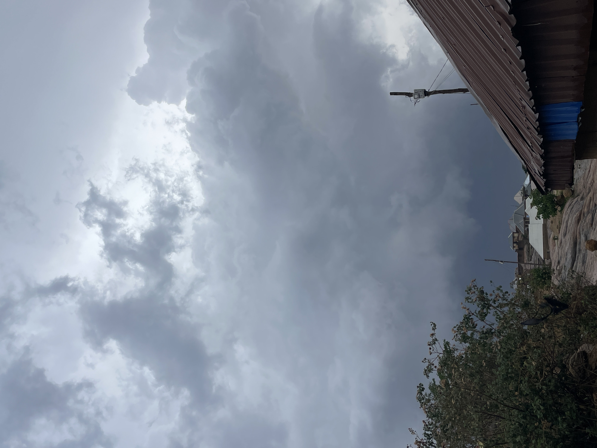 The image captures an overcast sky filled with dense, gray cumulonimbus clouds, indicative of a possible storm. In the foreground, a corrugated metal roof of a building is visible in the upper right corner. A utility pole and electrical box are positioned near the roof. Adjacent to the roof, the landscape descends to reveal a cluster of houses. A tree with green foliage is situated at the bottom right corner. The photo was taken in the city of Jos, Nigeria.