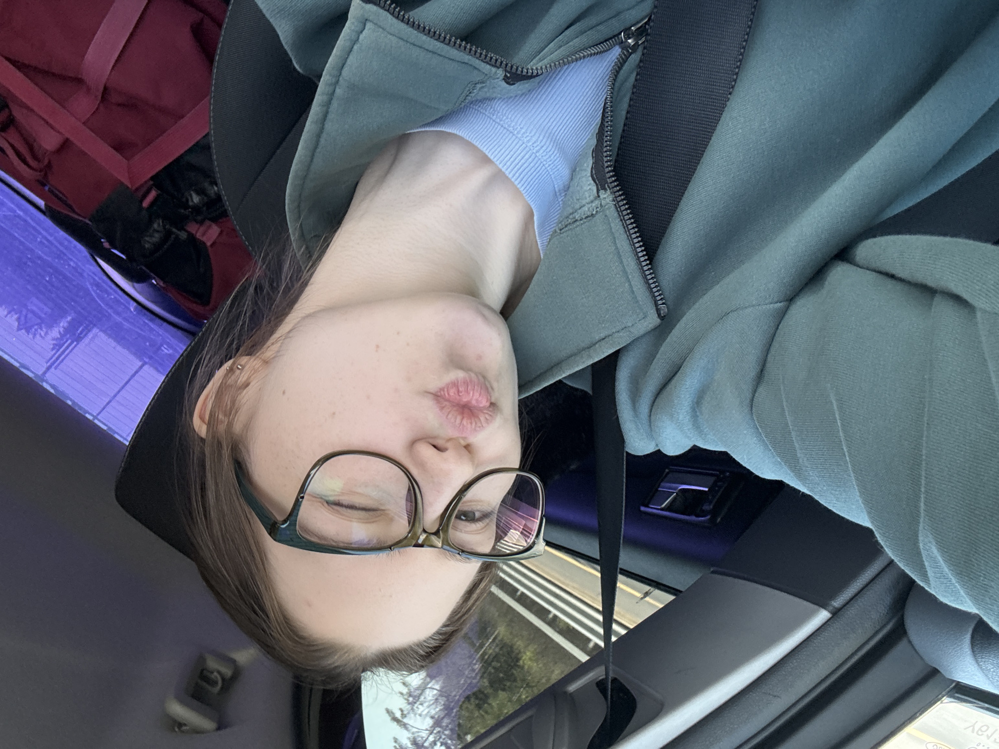 A young woman is seated in the driver's seat of a car, presumably traveling along a road near Pesochny, Russia. She is angled sideways, facing the camera with her head tilted. She wears glasses and a light-blue top, zipped into a green hoodie. A dark-colored seatbelt crosses her body. A red bag is positioned behind her. The background shows blurred trees and road markings visible through the car window, suggesting movement.