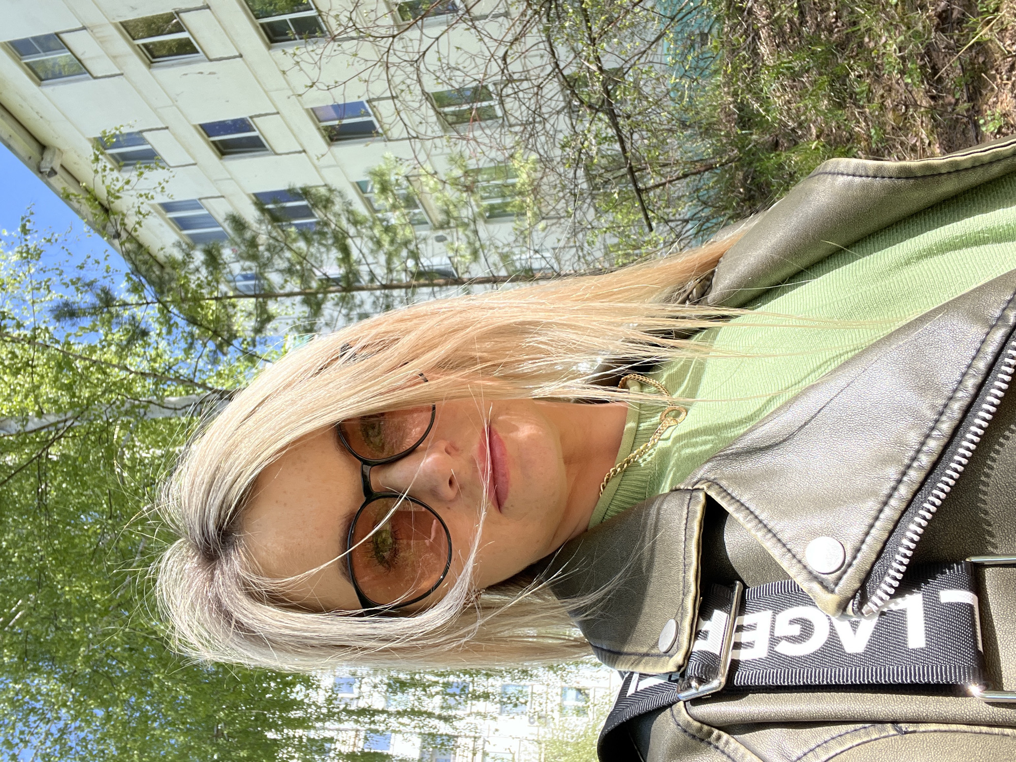 A woman with blonde hair and round sunglasses poses in an outdoor setting. She wears a light green sweater and a gray-green leather jacket with visible "LAGE" text on a black and white strap. Behind her are deciduous trees and a tall building with multiple windows under a blue sky, suggesting an urban landscape. This image was captured in the city of Chaikovsky, Russia.