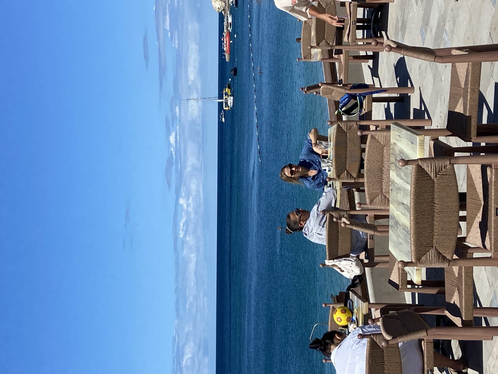 The image depicts an outdoor seating area at Petrovac centar, Montenegro, with tables and chairs set up facing the Adriatic Sea. Two women are seated at a table in the foreground, with one wearing sunglasses and a denim shirt, and the other facing away from the camera in a striped shirt. A yellow soccer ball is visible on a table to the left, near a person in a white shirt. In the background, the sea is calm with several boats and yachts anchored or sailing. A rope barrier with buoys marks a section of the water. The sky is bright blue with scattered white clouds. The scene conveys a relaxed atmosphere characteristic of a coastal town.