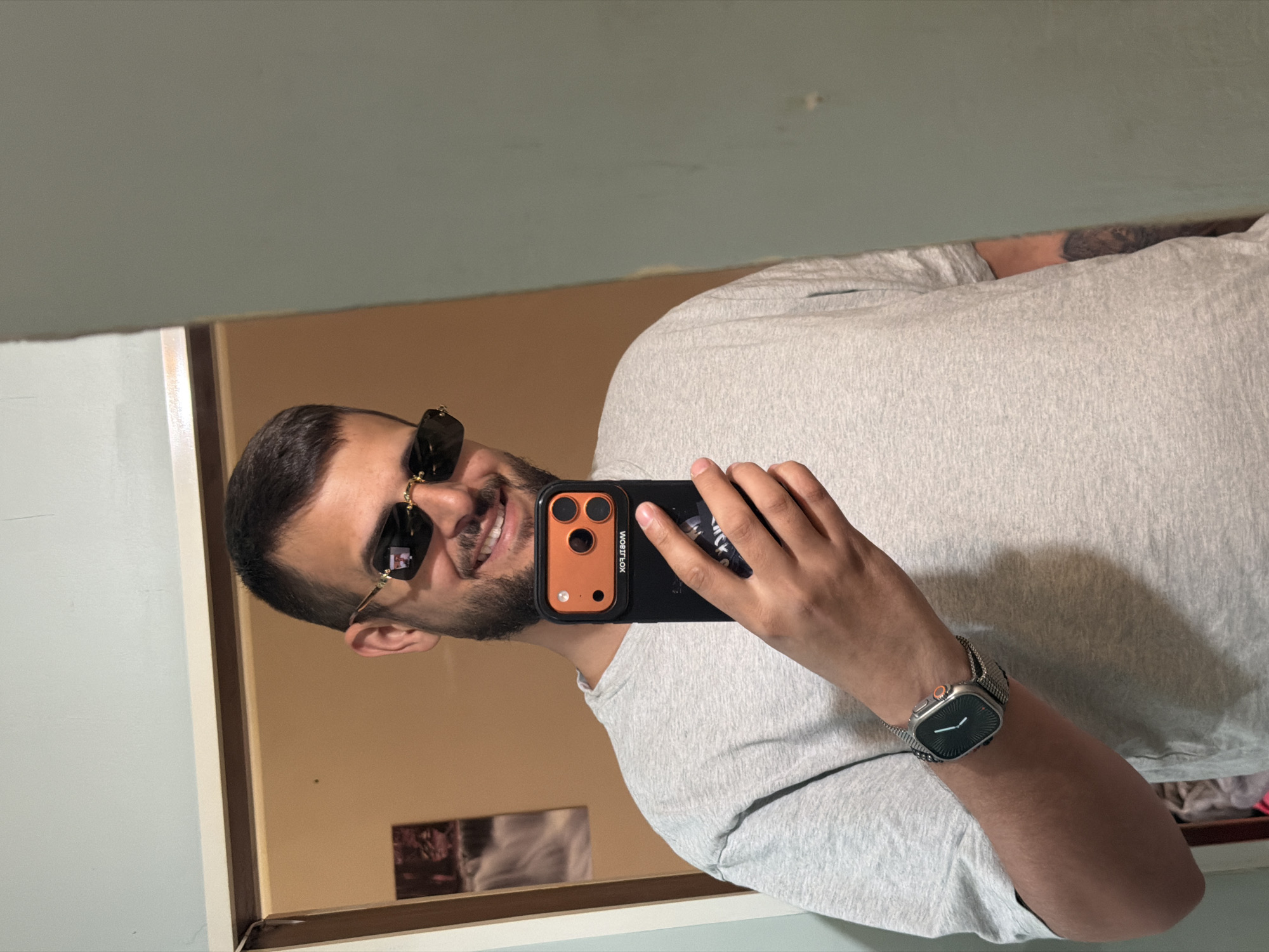 A man is taking a selfie in a mirror. The man is wearing sunglasses, a light gray t-shirt, and a watch with a silver band. He is holding a black smartphone with an orange camera housing. The background appears to be a room with light-colored walls and a mirror frame. There is no information to indicate this photo was taken in Sofia, Bulgaria.