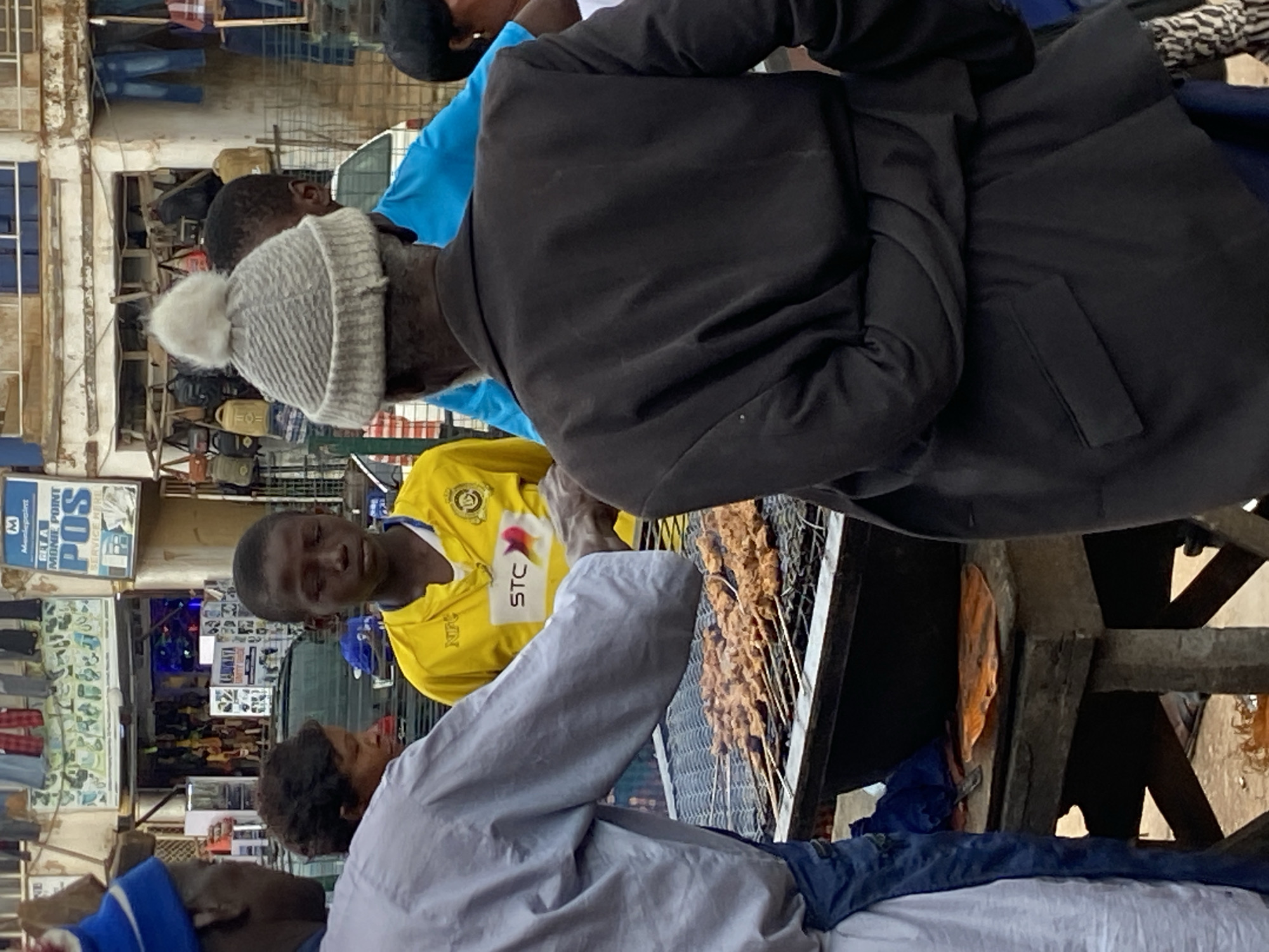 The image depicts an outdoor scene in the city of Bukuru, Nigeria, likely a market or street vendor area. In the foreground, a person wearing a blue hat and a light-colored robe is visible from the side, with their back to the camera. They appear to be tending to a grill laden with skewers of meat, suggesting a food preparation or sales activity. To their right, a young boy in a bright yellow shirt with "STC" prominently displayed is looking downwards. Behind him, another individual in a blue shirt is partially visible. Further back, an older person in a dark jacket and a knitted hat with a pom-pom is seen from the back, facing the grill. The background reveals a bustling environment with shops, hanging merchandise, and a sign that reads "MEET POINT" and "POS SERVICE HERE." The overall impression is a lively street scene in Nigeria, focusing on a food vendor preparing a meal.