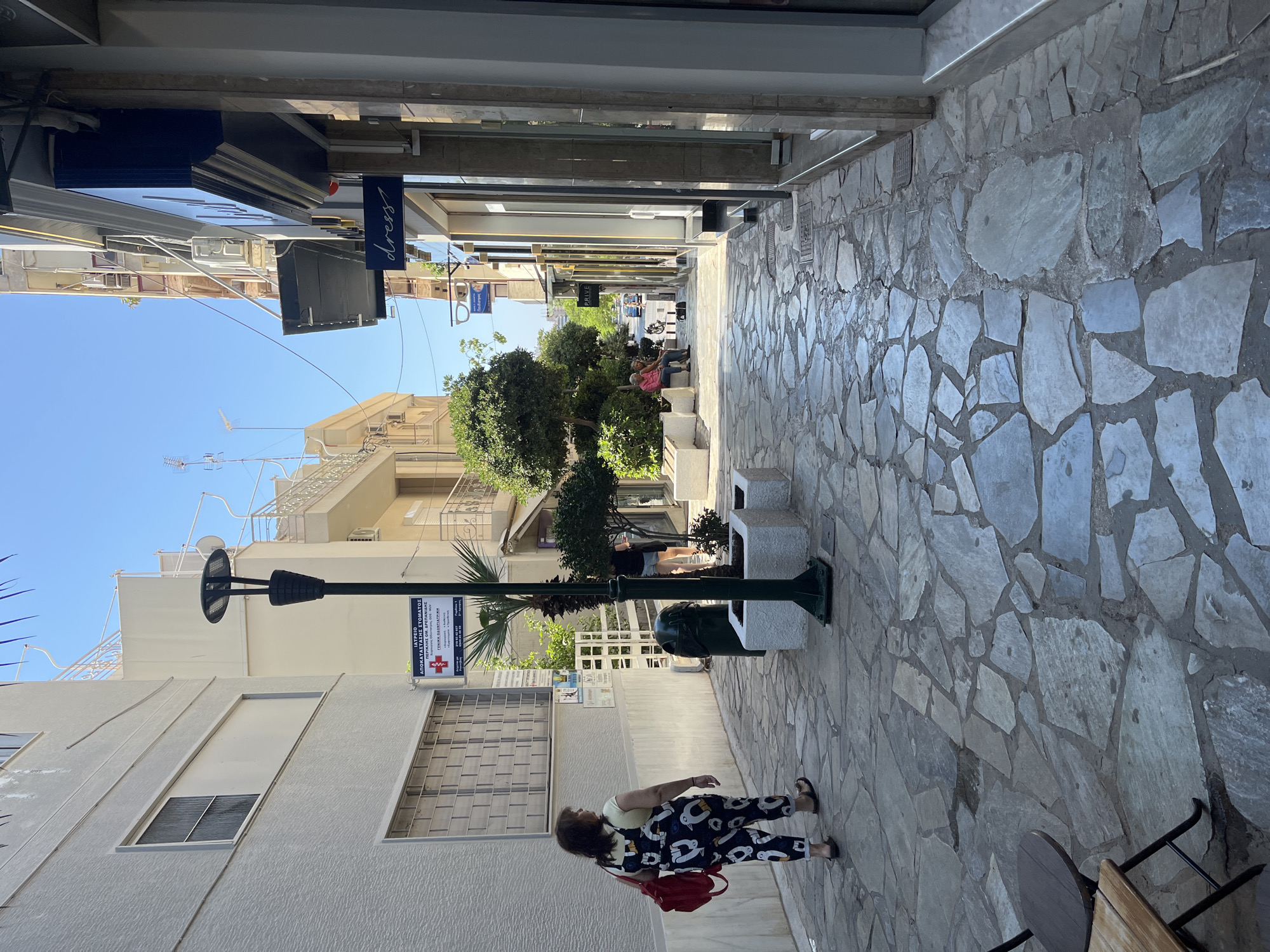 A woman in a white shirt and black overalls with a white penguin print walks away from the camera down a narrow cobblestone street in Nea Smirni, Greece. She carries a red backpack on her left shoulder. The street is lined with buildings on both sides. On the left, a light-colored building has several windows with metal grates. A medical sign is posted on the wall. A tall, dark lamppost with a modern LED fixture stands in the middle of the street. To the right of the lamppost, there is a dark green trash can next to a white planter with a small palm tree. Further down the street, on the right, are shops with large glass windows and awnings. One shop has a sign that reads "dress" in white script. Across the street, there are more trees and people sitting on benches. The sky is bright blue with no clouds. The sun is high, indicating it is midday.