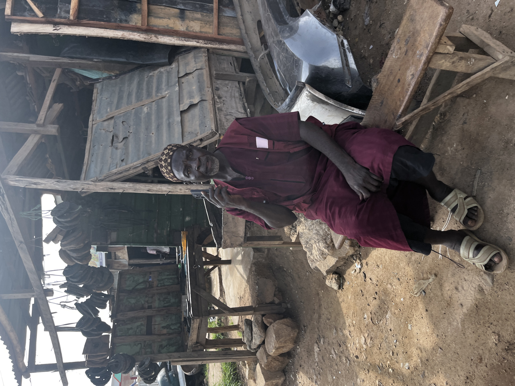A middle-aged African man with dark skin, a beard, and a patterned cap sits on a rustic wooden bench in front of a makeshift structure in Jos, Nigeria. He wears a maroon tunic and black trousers, with his feet clad in light-colored sandals. The man is holding a smartphone to his ear, suggesting he is engaged in a conversation.
The environment appears to be an outdoor marketplace or workshop. The structure behind him is constructed from corrugated metal and rough-hewn wood, with various items hanging from the roof, possibly parts or tools. In the foreground and to the right, there are car parts, specifically what appears to be a bumper, scattered on the ground. The ground is a mix of dirt and sand, with some stones and debris. The overall impression is of a busy, somewhat cluttered, and utilitarian space.