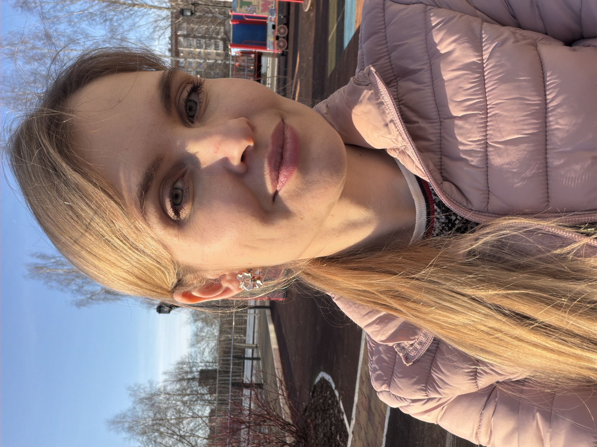 A woman is pictured outdoors. She wears a pink puffer jacket and has blonde hair. In the background, a children's play area is visible, including a red train-themed play structure. Trees are also present, and the sky is clear and blue. Based on provided context, the location is Izhevsk, Russia.
