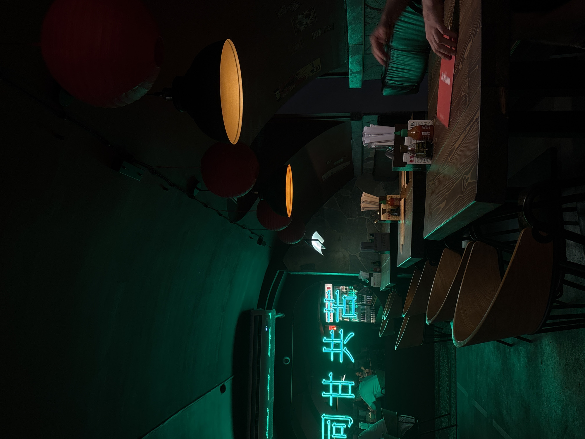 A dimly lit, unoccupied dining area is visible, with a green-tinged lighting scheme. The location is Perm, Russia, indicated by the menu on the table and bottles of what appears to be Hong Kong sauce and Kikkoman soy sauce. The image shows a narrow space with chairs set up along a wall, suggesting a restaurant or bar setting. Japanese or Chinese characters are displayed in neon signs. Red lanterns and pendant lights provide illumination. Chopsticks and condiment containers on the tables indicate a focus on Asian cuisine. There's a partially visible person in a green shirt and an individual's hands placing a menu. The environment is dark, with the green and yellow light creating a distinctive ambiance.