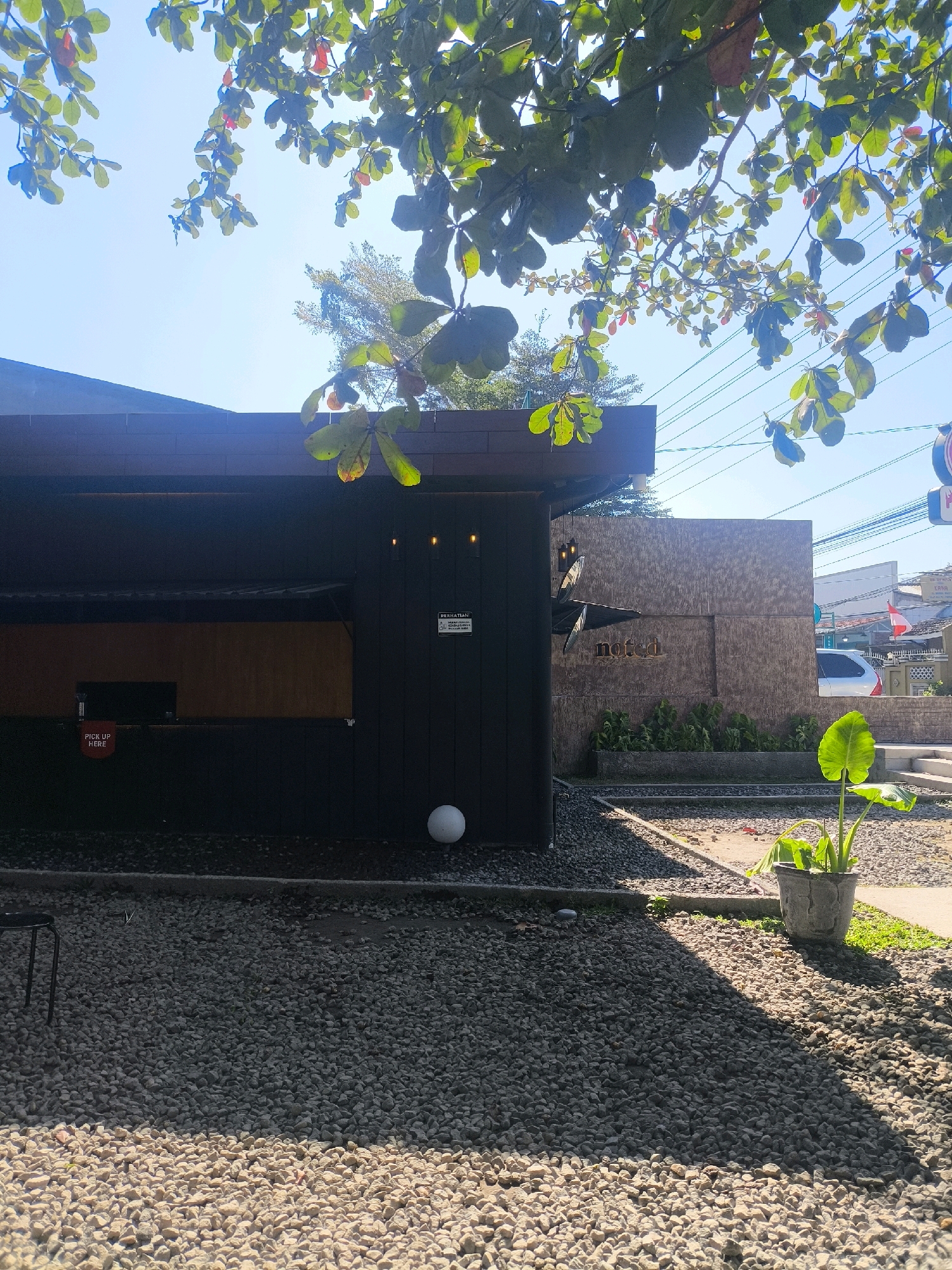 The image captures a view of a contemporary outdoor establishment, likely a cafe or restaurant, situated in Ngaglik, Indonesia. The structure features a dark facade with a visible counter area and a sign indicating "PICK UP HERE." A white spherical object rests on the gravel ground near the building. To the right, the establishment displays signage with the word "notched." The surrounding area is covered in gravel, with a potted plant featuring large green leaves in the foreground. Overhead, tree branches with green and reddish leaves are visible against a clear blue sky. Utility lines are also present in the background. The scene appears to be set during the daytime under sunny conditions.