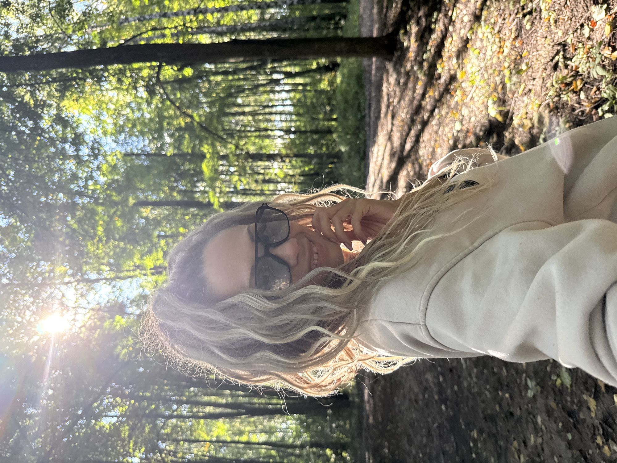 In Rusanovo, Russia, a woman with blonde, curly hair pulled into a high ponytail is captured in a selfie taken outdoors in a sun-dappled forest. She is wearing dark sunglasses and a light-colored sweatshirt. Her right hand is raised to her face, with her fingers gently touching her cheek. The background is filled with tall trees, their leaves illuminated by the bright sunlight filtering through the canopy, creating a bokeh effect. Shadows from the trees are cast across the forest floor, which is covered with fallen leaves and some green undergrowth. The scene appears to be a pleasant, sunny day in the woods.