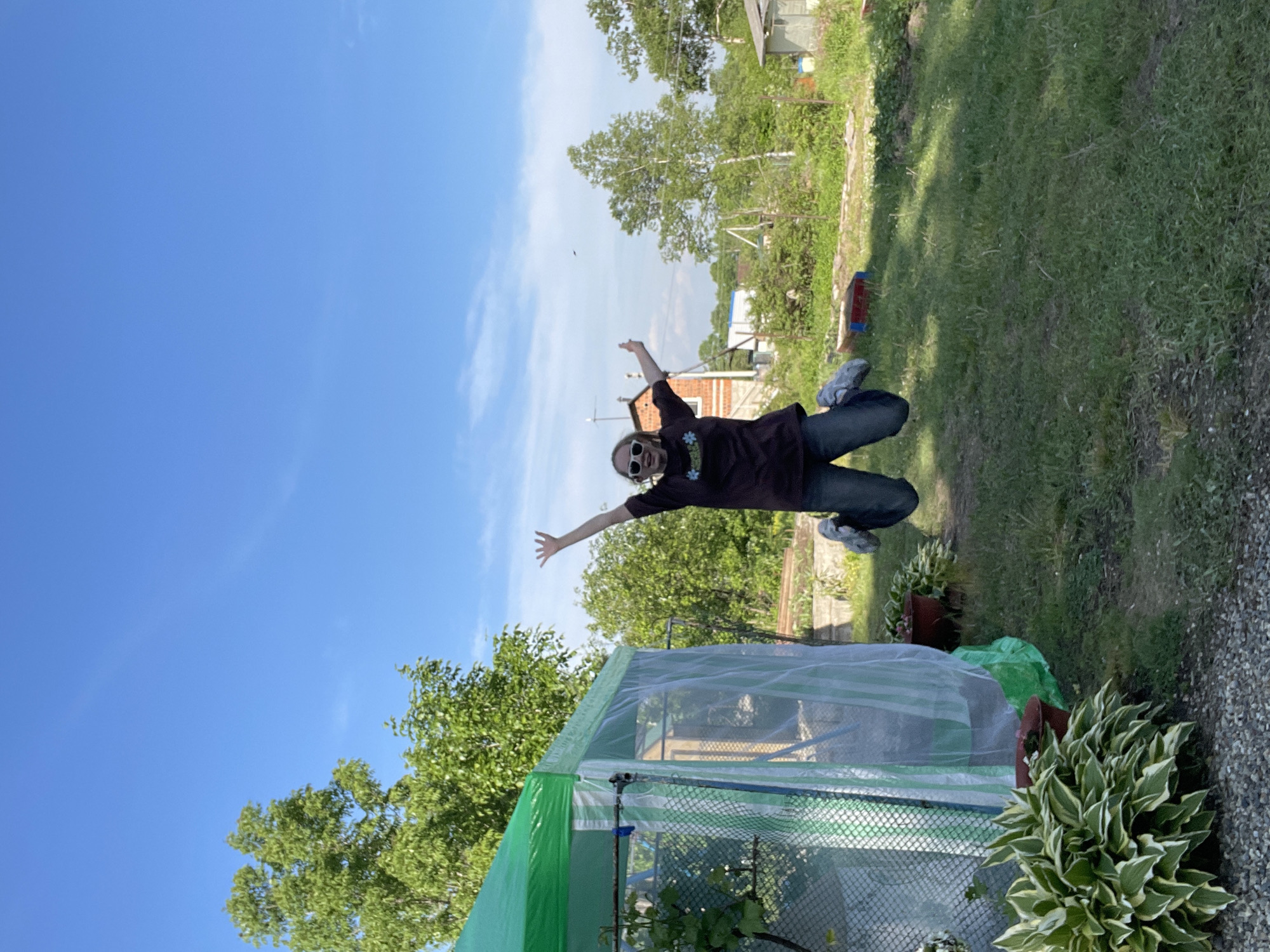 A person wearing dark clothing, jeans, and sunglasses is jumping in the air with arms outstretched. The background features a green lawn, trees, a blue sky with some clouds, and a structure covered in a translucent material. Based on the presence of characteristic garden elements and the overall scenery, the location is likely a backyard or garden area. I cannot confidently determine that this image was taken in the city of Yasnoe, Russia.