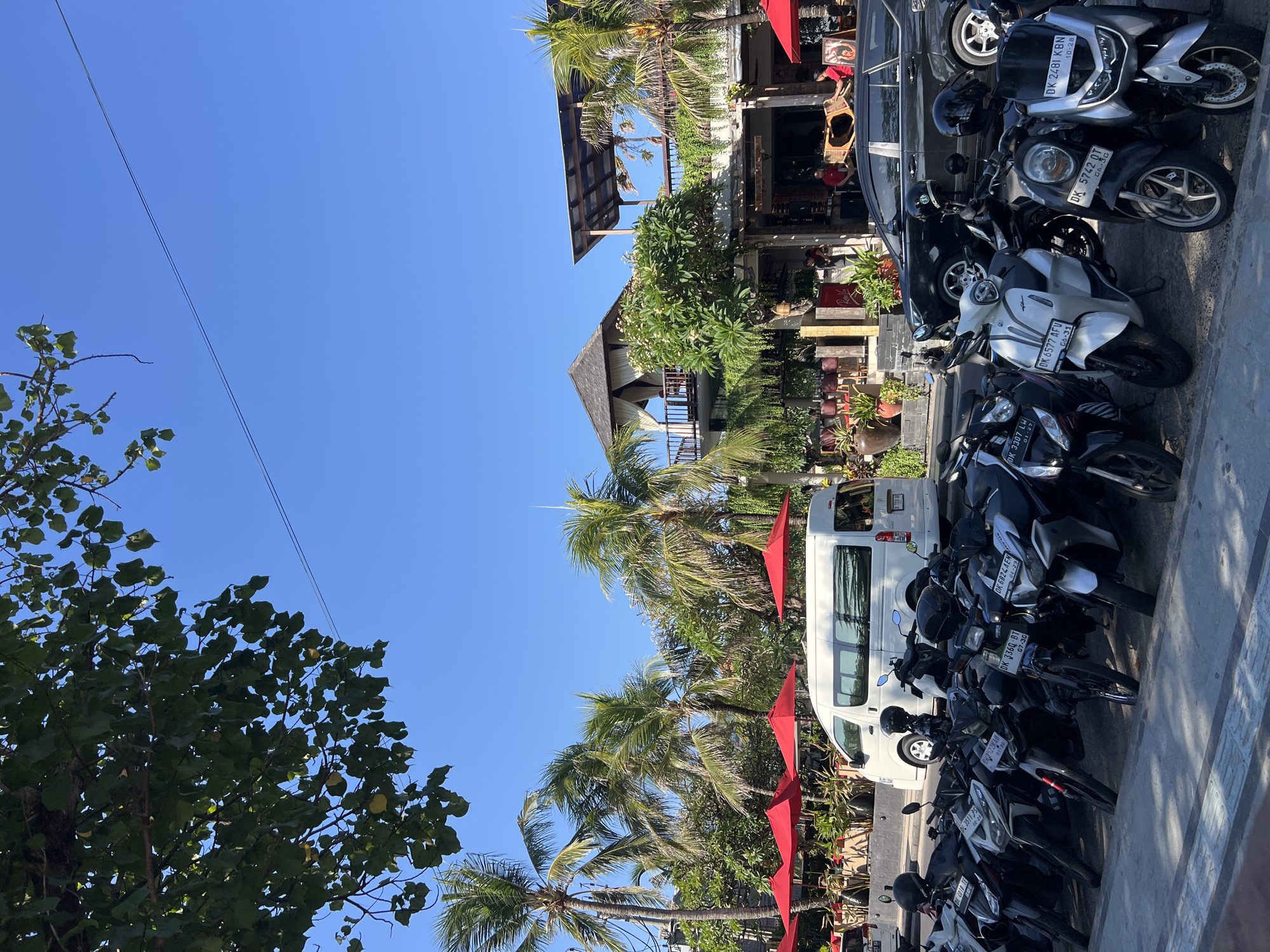 The image depicts a sunny day on a street in Kuta, Indonesia, likely near a beachside establishment. In the foreground, a row of parked motorcycles, predominantly scooters, are lined up along the curb. Their license plates are visible, with some displaying the "DK" prefix common in Bali. Behind the motorcycles, a white van is parked. Further back, a building with multiple levels features balconies and open-air seating areas, suggesting a restaurant or cafe. Red umbrellas are set up outside some of these seating areas. Palm trees and lush greenery are abundant throughout the scene, indicative of a tropical environment. Several people are visible, some sitting at the outdoor tables and others walking near the building. A black car is also parked on the street. The sky is clear and blue. The overall impression is one of a vibrant, tropical, and bustling tourist area.