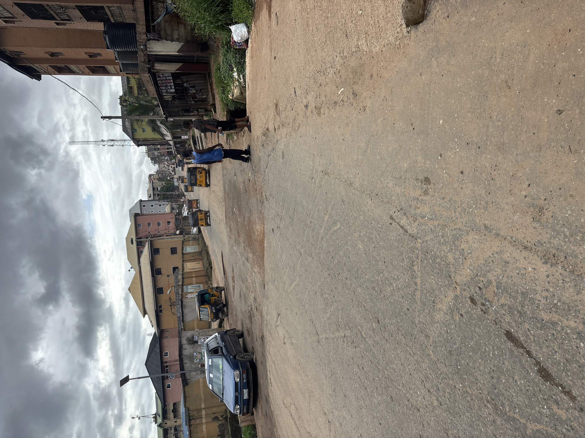 The image depicts a street scene in Nkpor, Nigeria, under a cloudy sky. The road is paved but shows signs of wear and tear, with visible potholes and patches of dirt.
On the left side of the road, a blue Volkswagen Golf car is parked. Further down the road, several yellow auto-rickshaws, commonly known as "Keke Napeps," are present, some in motion and others parked.
Two individuals, a man in a blue t-shirt and dark trousers and another person in a patterned top and dark trousers, are standing in the middle of the road, seemingly engaged in conversation.
The background is lined with buildings of various sizes and architectural styles, some appearing to be multi-story residential or commercial structures. A telecommunication tower is visible in the distance. Greenery, including grass and some small trees, is present along the sides of the road and in patches between buildings. A white bag is visible on the right side of the road, near some dense vegetation. The overall atmosphere suggests a bustling urban environment with moderate activity.