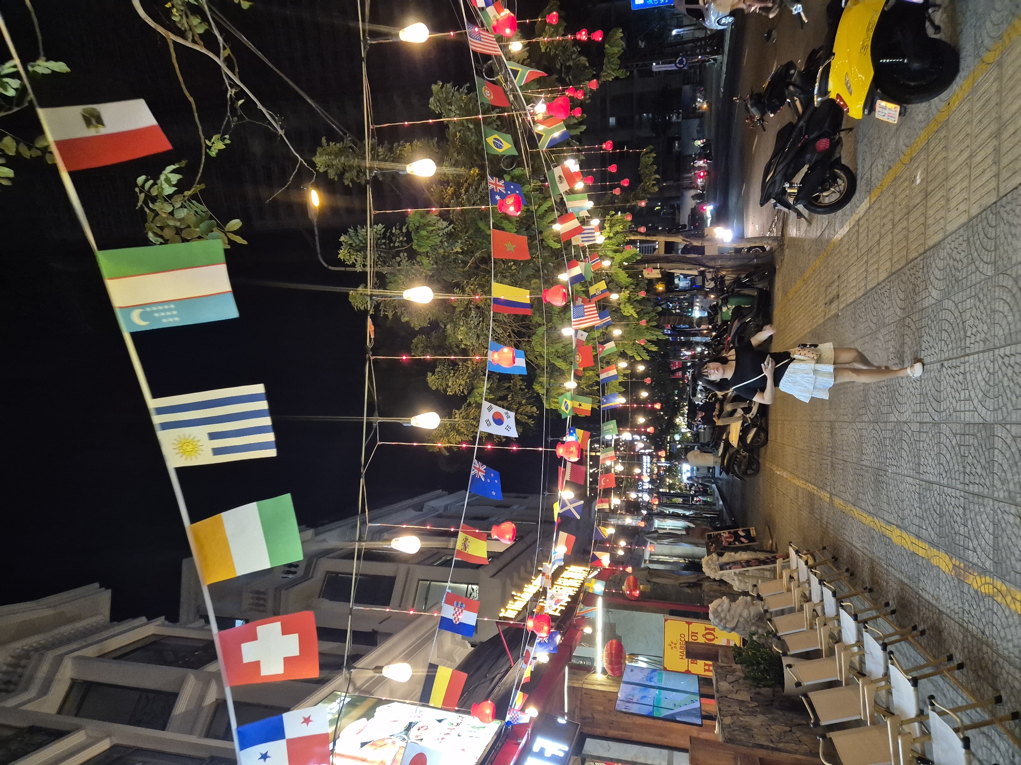 The image depicts a street scene at night in Da Nang, Vietnam, adorned with numerous national flags strung across the street and interspersed with decorative lights. A young woman wearing a black top and a white skirt is walking down the sidewalk, carrying a bag over her shoulder. The sidewalk is lined with empty chairs, likely for outdoor seating at nearby establishments. Several motorcycles are parked on the street. The architecture suggests a commercial area, with buildings and shopfronts visible in the background. The atmosphere is festive and lively, indicated by the flags and lights.