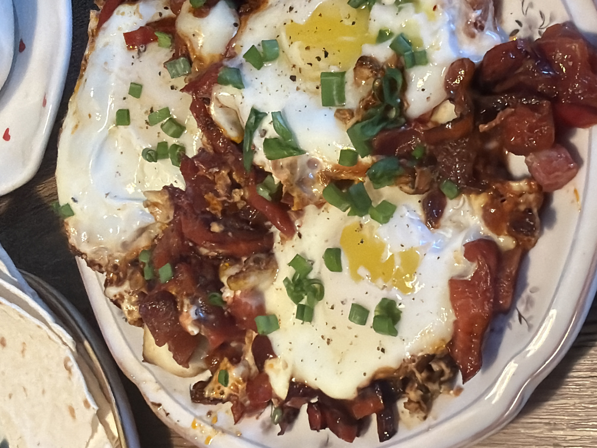 The image depicts a plate of food consisting of fried eggs, roasted red peppers, and chopped green onions. The eggs are cooked sunny-side up, with the yolks visible. The roasted red peppers appear to be sautéed with some type of sauce. Chopped green onions are scattered on top as a garnish. A stack of tortillas is partially visible in the lower left corner of the image.