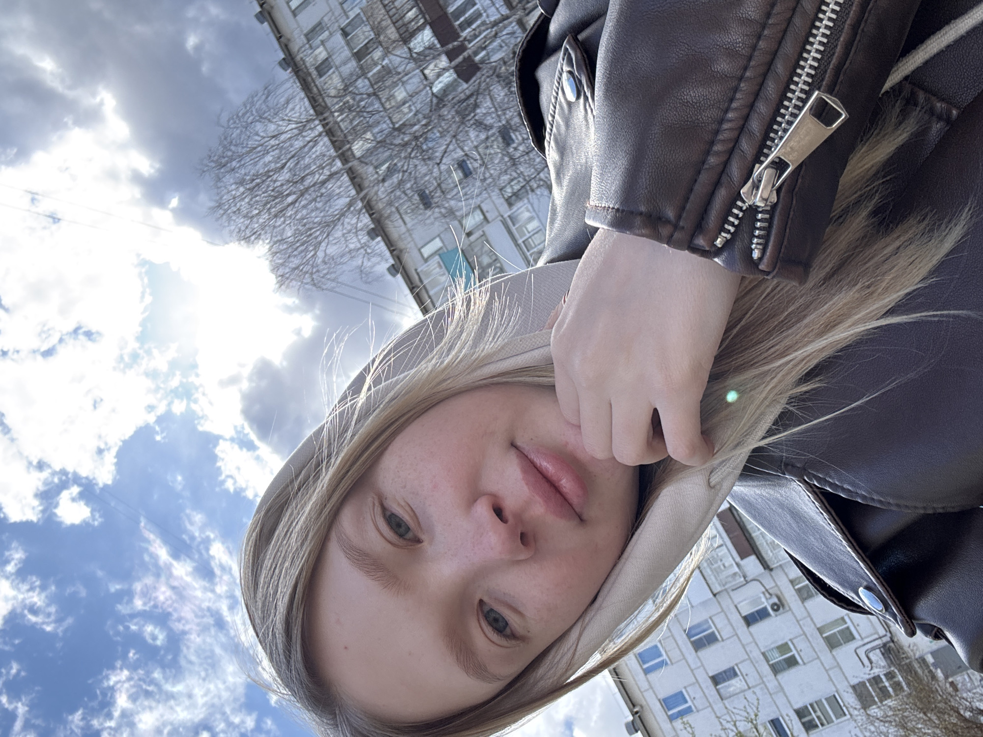 An upward-angled outdoor selfie captures a young woman in Chaikovsky, Russia. She has blonde hair, light-colored eyes, and fair skin with visible freckles, looking directly at the camera with a neutral, relaxed expression and slightly parted, full lips. She wears a light-colored hooded top beneath a dark brown or black leather-like jacket, which features a prominent silver zipper on the sleeve.
The setting is an urban environment under bright daylight. The sky is partially blue with significant white and grey cloud cover, and the sun is visible in the upper left, casting a strong glare. In the background, a tall, multi-story residential building with many windows and external air conditioning units is visible. Bare tree branches are scattered throughout the background, suggesting a late winter or early spring season. No visible text is present.
