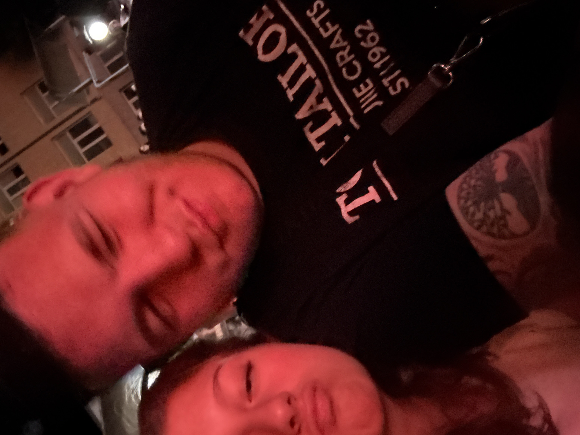 A selfie taken in low lighting conditions with a red hue. Two people are visible in the frame. The person in the foreground on the right is a male with short hair, appearing to be looking down and to the left, with his eyes closed. He is wearing a black t-shirt with white lettering that reads "TAILOR" and below it "THE CRAFTS 1962". He has a tattoo on his left forearm featuring a tree with roots. The person on the left appears to be a younger female, also looking towards the camera with her eyes partially closed and her lips pursed. She has long, dark hair. The background shows a building with windows and a street lamp, suggesting an outdoor urban setting at night.