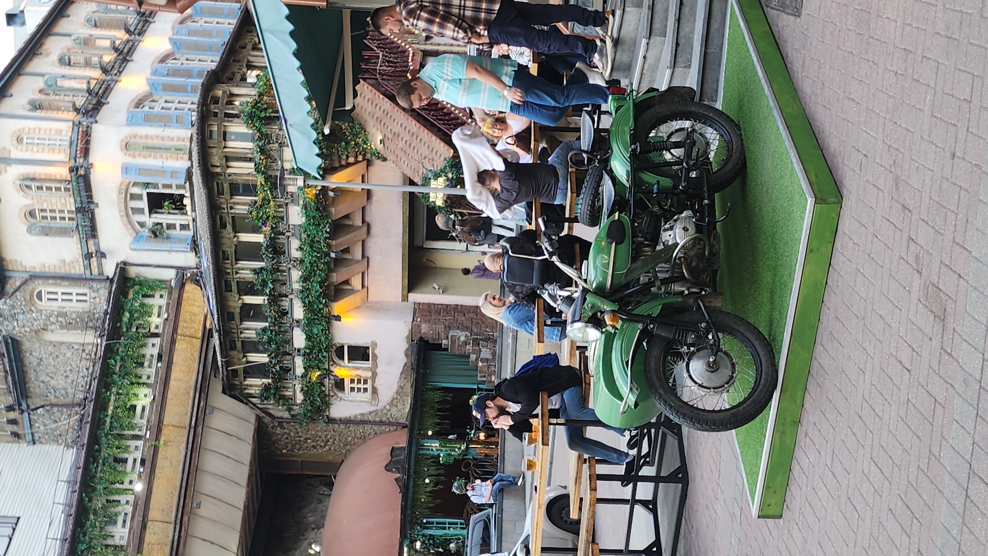 This is a medium-angle, eye-level shot of an outdoor seating area outside a building with a facade decorated with greenery. In the foreground, a green motorcycle with a sidecar is parked on a raised green platform. The motorcycle is the main subject of the image, positioned centrally. Behind the motorcycle, several people are seated at wooden picnic tables, some appearing to be dining or drinking. The building in the background features multiple stories with arched windows and a rustic stone and stucco exterior, with some windows shuttered with blue. The overall atmosphere suggests a casual outdoor dining or social gathering space in an urban setting, possibly in a historic or themed district. The pavement in the foreground is brick-laid. The sky is not visible, suggesting the photograph was taken during daylight hours.