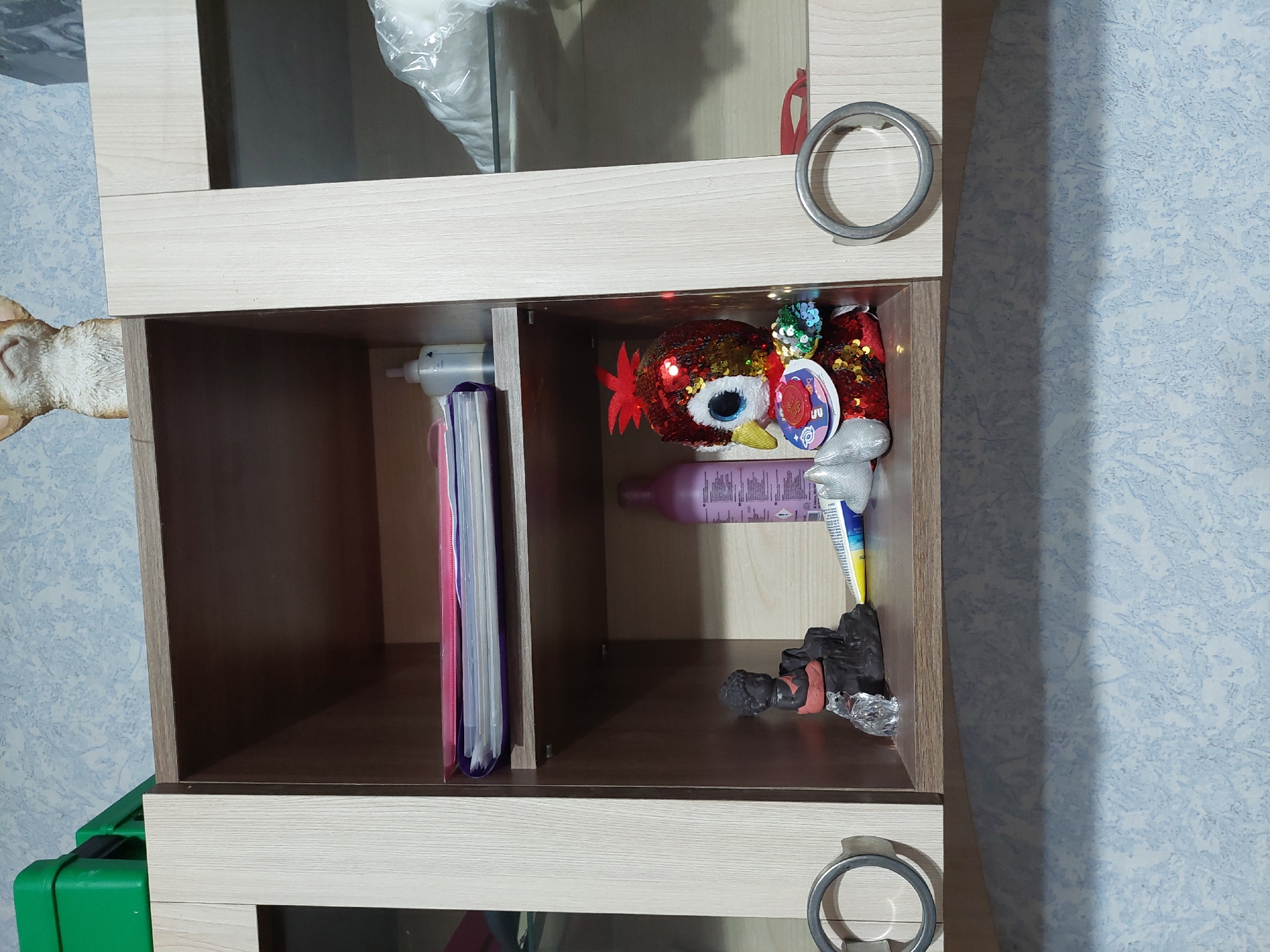 The image depicts an interior scene, specifically a section of a room in the city of Orhei, Moldova. A wall-mounted, three-section cabinet unit is centrally positioned. The two outer sections feature light wood-grain doors with clear glass inserts, each fitted with a circular, metallic gray handle on the lower corner. The central section is an open shelving unit, matching the light wood-grain finish.
The top shelf of the open unit holds a stack of binders or folders, primarily pink, purple, and white, alongside a white pump-top dispenser bottle on the right. The bottom shelf contains diverse objects: on the left, a dark gray figurine of a meditating figure with an orange sash, resting on a black, textured base. A small, clear, crystalline animal figurine (possibly a cat) sits in front of it. Towards the center, a yellow and blue tube (e.g., toothpaste or cream) lies horizontally, next to a small, crinkled silver object. On the right, a plush toy bird, approximately 20cm tall, is covered in red and gold sequins, featuring large blue eyes, a yellow beak, and a red crest. A circular tag with red and blue designs is attached to its chest. A tall, light purple plastic bottle with an unreadable label stands behind the plush toy.
Above the cabinet, a light brown ceramic or resin figurine of an animal's head (likely a dog or cat) is partially visible at the top center, and two stacked green plastic containers are present on the upper left. The wall behind and below the cabinet is covered in light blueish-gray wallpaper with a subtle, abstract lighter pattern.