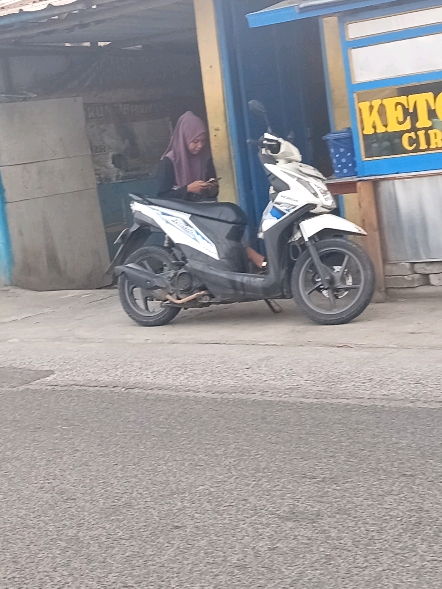 A white and black scooter is parked on a paved surface in front of a small building. A woman wearing a purple hijab and black clothing is seated on the scooter, looking at her phone. The building has blue and yellow accents, and a sign with yellow lettering that appears to say "KETOK" is visible. There is a blue plastic container on a shelf outside the building. The ground in front of the building is a light gray paved surface, and the street in the foreground is a darker gray asphalt. The background shows a corrugated metal roof over the building and a wall with some posters on it.