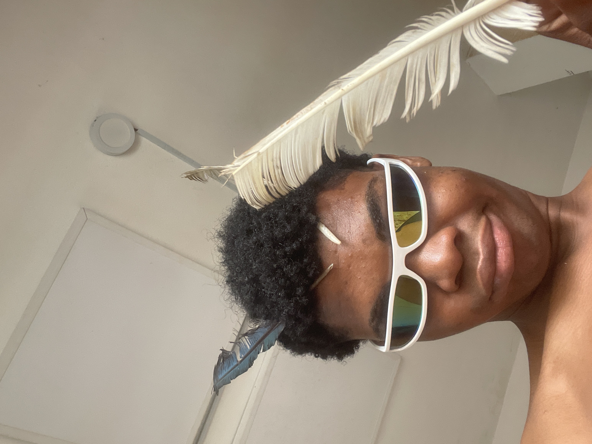 A close-up, low-angle shot shows a Black person wearing white, futuristic sunglasses with reflective lenses. The person has dark, curly hair with two feathers tucked into it: one blue feather on the left side of their head and a large, off-white feather held in their right hand, positioned near their face on the right side. Another smaller, light-colored feather is visible sticking out from the person's forehead. The background consists of a plain, light-colored wall and ceiling with a circular light fixture. The image appears to be taken indoors in a well-lit room. There is no discernible indication of the location being in Ado, Nigeria.