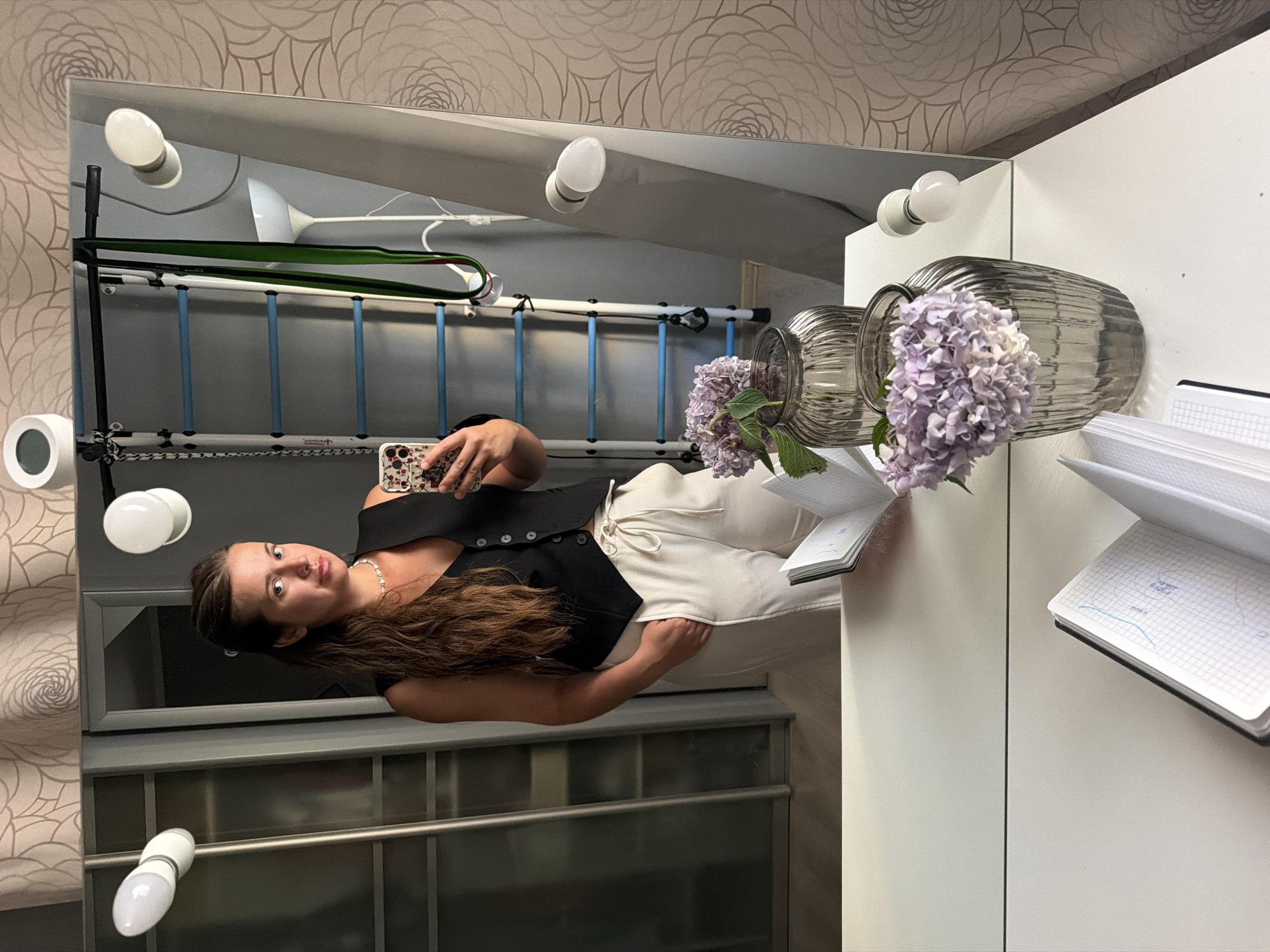 The image depicts a young woman with long brown hair, wearing a black vest and cream-colored pants, standing in front of a mirror. She is holding a smartphone in her right hand and has her left hand in her pocket. In the reflection of the mirror, a home gym apparatus is visible, along with light fixtures and wallpaper with a spiral pattern. On the table in front of the mirror, there are two glass vases with purple hydrangeas and an open notebook. The scene appears to be indoors, likely in a residential setting. The city of Sochi, Russia is not depicted in the image.
