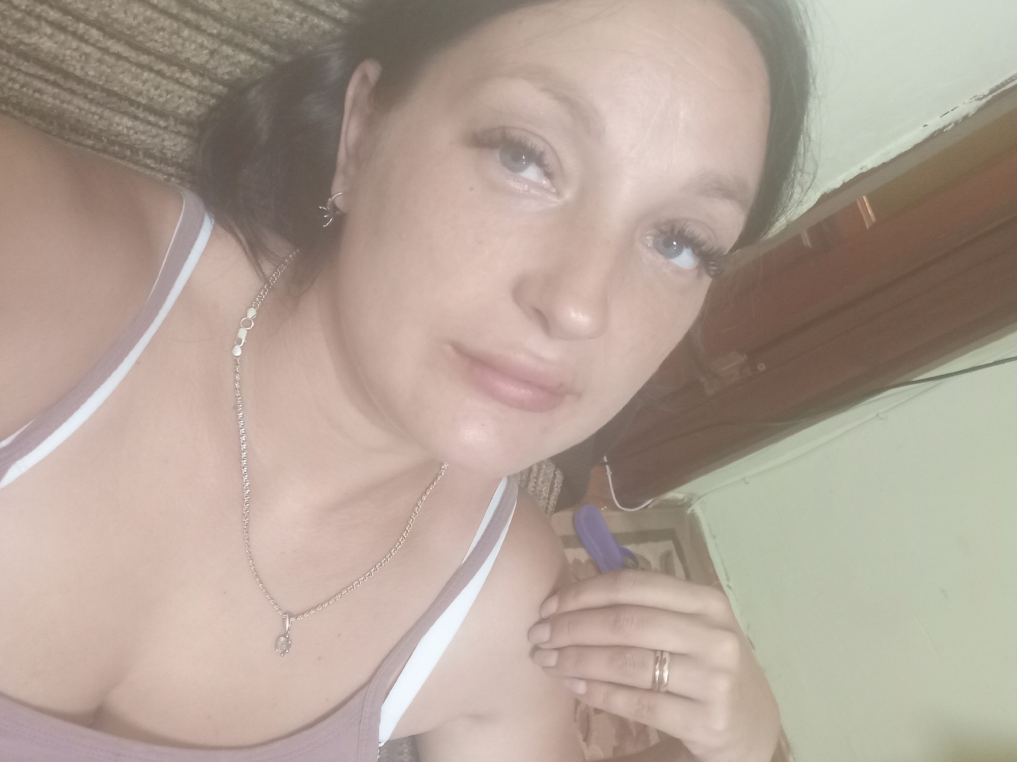 The image shows a close-up, overhead shot of a young woman lying down. She has light skin, blue eyes, and long, dark eyelashes. She is wearing a light-colored tank top with white straps and a thin silver necklace with a small pendant. A ring is visible on her right hand. The background is out of focus but appears to be an indoor setting with neutral-colored walls and furniture. There are no discernible landmarks or specific environmental details that would indicate a location in Chuhuiv, Ukraine. The image is a selfie, with the camera angled slightly downwards.