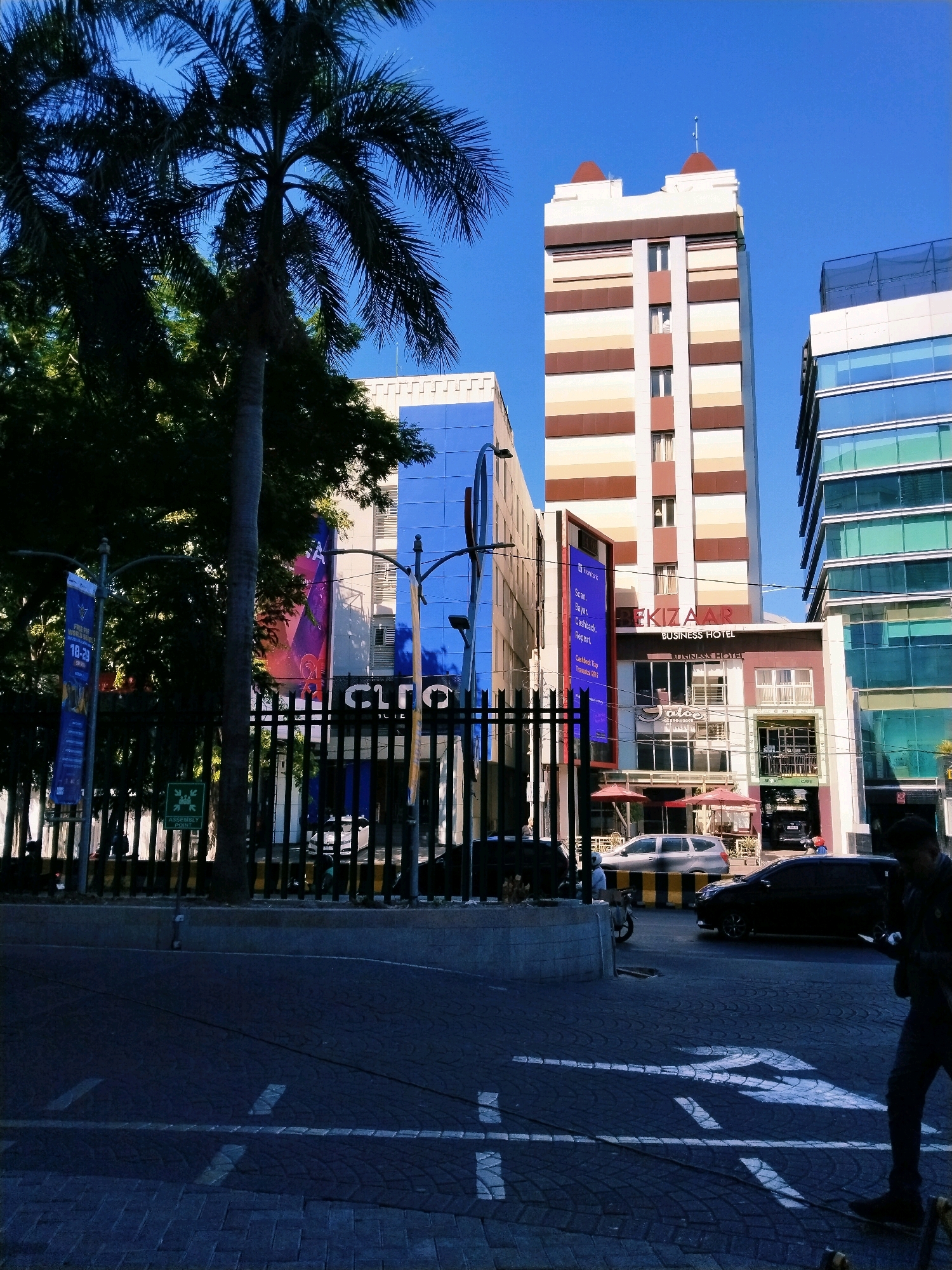 The image captures a street scene in Surabaya, Indonesia, on a clear day with a bright blue sky. In the foreground, a dark asphalt road with patterned paving stones is visible, marked with white directional arrows indicating traffic flow. A person in dark clothing is walking on the right side of the frame, looking down at a device in their hands.
Dominating the background are several modern buildings. To the left, a tall palm tree stands in front of lush green foliage. Behind the greenery, a building with a vibrant blue facade is partially visible. Adjacent to it is a tall, slender tower with alternating bands of cream and brown, topped with decorative finials. To the right of this tower is another modern building with a facade of blue-tinted glass and white horizontal accents.
Several cars are present on the road, including a silver SUV and a dark sedan. A motorcycle is also parked near the fence. The buildings display various signage. One prominent sign reads "EKIZAA BUSINESS HOTEL". Another sign on a blue banner has text that appears to be an advertisement. A green sign indicating an "ASSEMBLY POINT" is affixed to a pole near the fence. The area is secured by a black metal fence. The scene suggests a commercial or business district in the city.