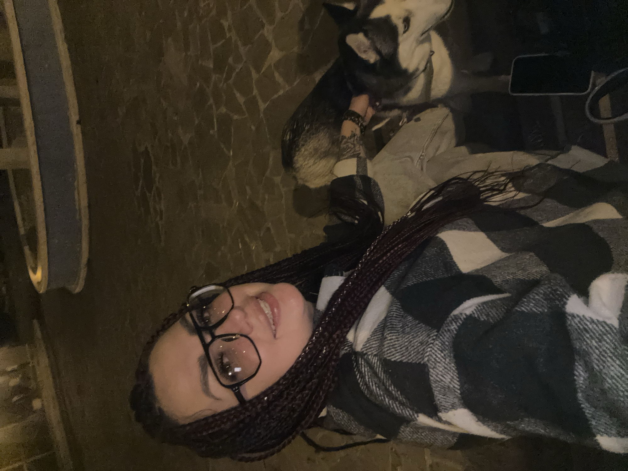 A young woman with braided dark hair, wearing large black-rimmed glasses and a black and white checkered jacket, is taking a selfie outdoors at night. She is smiling and looking at the camera. A husky dog is next to her, its head resting on her lap. They are on a cobblestone surface, and in the background, there is a concrete structure with a blue band around it. A dark object, possibly a phone, is visible on the ground to the right of the woman. The lighting suggests it is nighttime, and the environment appears to be an urban setting. The specific location in Tolyatti, Russia, cannot be determined from the visual information provided.