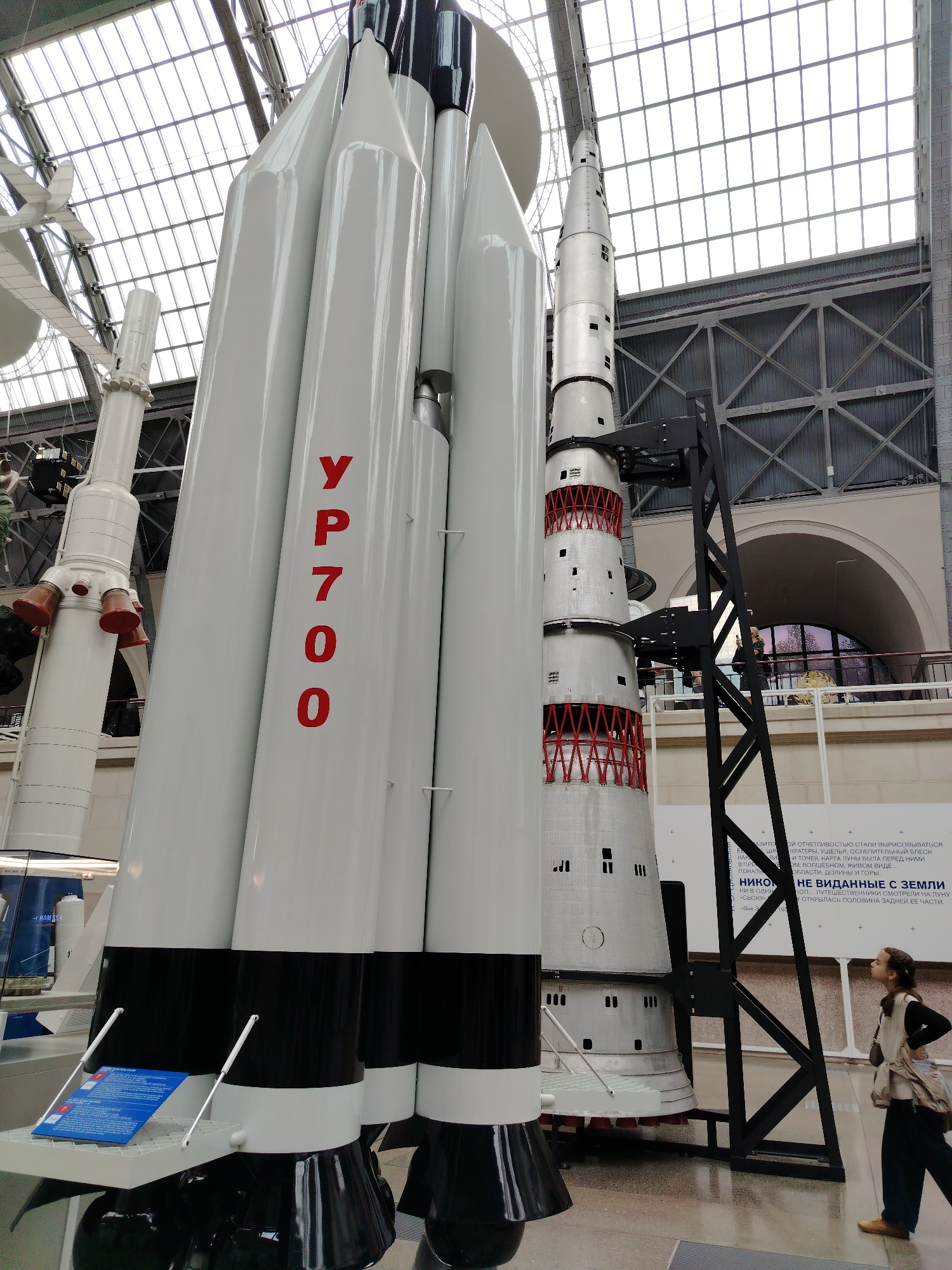 In Moscow, Russia, at the State Museum of Cosmonautics, a large rocket model is on display indoors under a glass and metal ceiling. The rocket is white with black and white striped lower sections. Red lettering on the side of one of the rocket stages reads "УР 700". A person is visible in the lower right corner, looking up at the rocket. They are wearing a dark vest over a lighter top, dark pants, and brown shoes. Another smaller rocket model is visible to the left of the main rocket. A black metal structure supports part of the larger rocket. Informational displays are present near the base of the rocket.