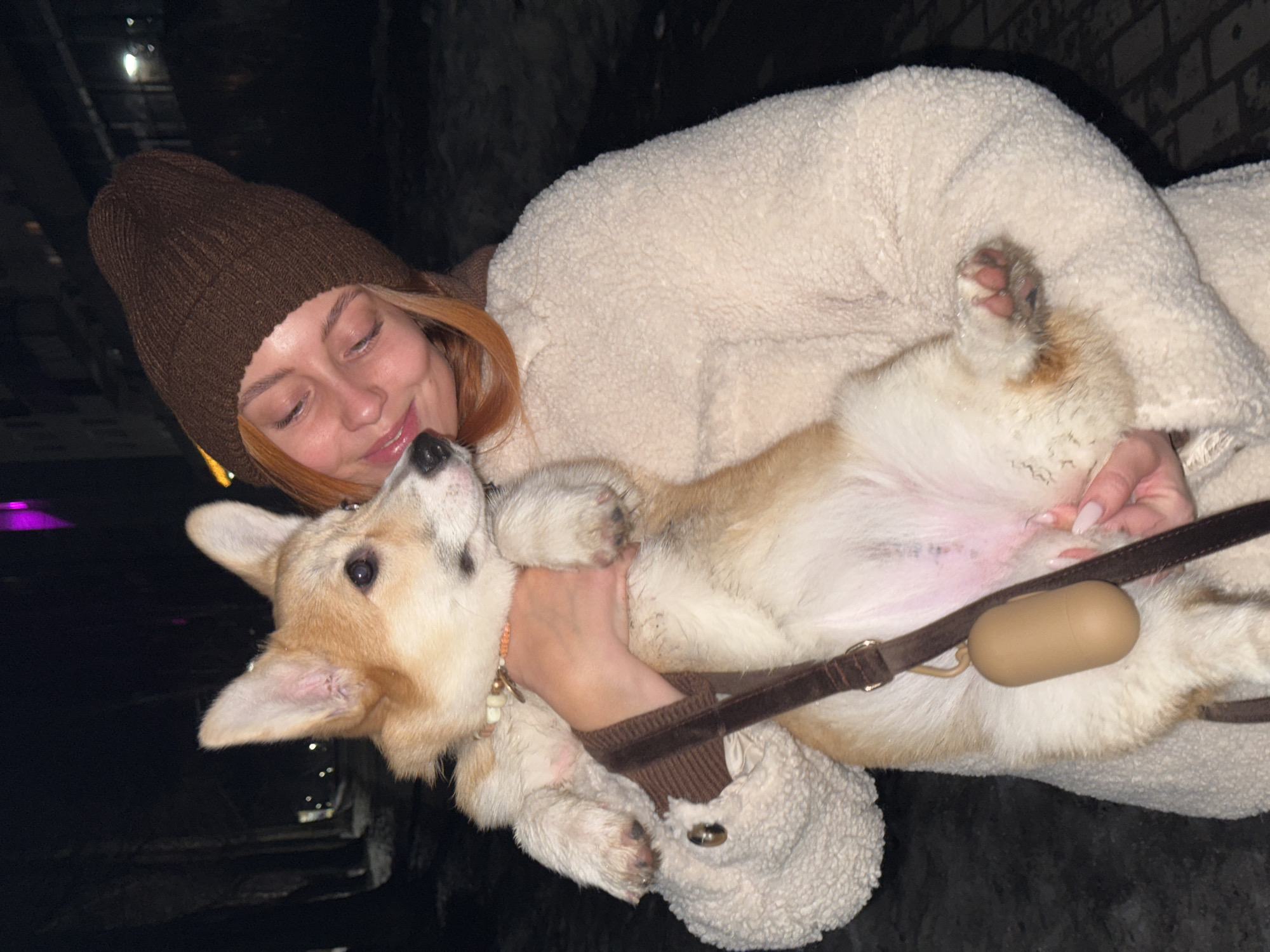 A young woman is holding a dog. The woman has red hair and is wearing a brown beanie and a cream-colored, textured coat. She is smiling and looking at the dog. The dog is a corgi, lying on its back. It has light brown and white fur, and is wearing a collar. There is a brown leash attached to the collar, with a brown capsule-shaped object attached to the leash. The background is dark and somewhat out of focus. It might be taken in the evening or at night. The location is Chaikovsky, Russia.