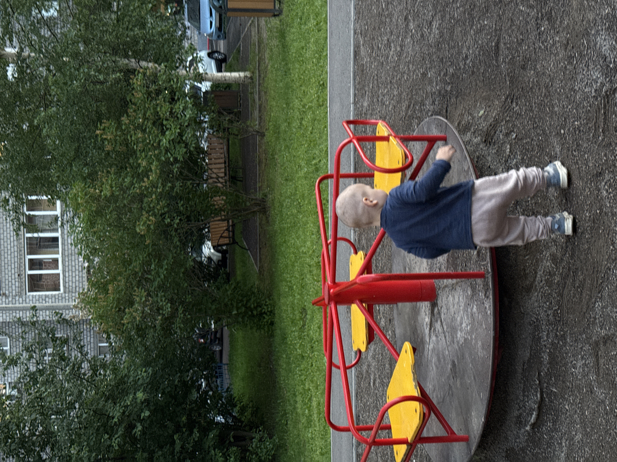 The image depicts a child's playground with a red merry-go-round featuring yellow seats. A young boy wearing a dark blue long-sleeved shirt and light-colored pants is standing near the merry-go-round, facing away from the camera. The playground is located in an urban environment, with a residential building visible in the background, characterized by gray brickwork and white-framed windows. Trees and green grass surround the playground. Several cars are parked in the background, and a wooden bench is also present. The overall scene suggests a typical playground setting within a residential area of Krasnoyarsk, Russia.