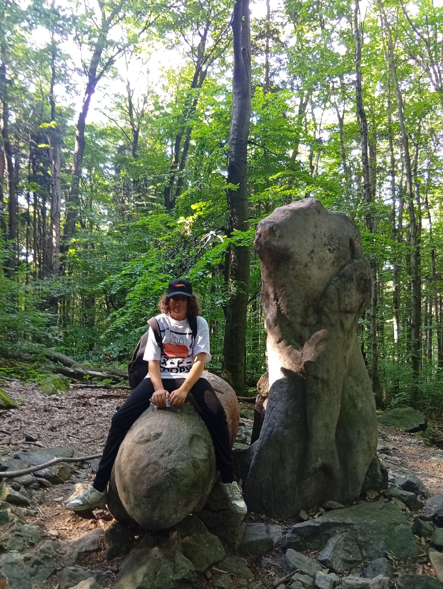 A person with medium-length curly hair, wearing a white t-shirt with an orange graphic, black pants, a black baseball cap, and sneakers, is sitting on a large round boulder in a forest. The person is looking towards the camera. To the right of the person is a tall, irregularly shaped rock formation. The ground is covered with rocks, dirt, and fallen leaves. Tall trees with green foliage form a canopy overhead, allowing sunlight to filter through. The scene appears to be the "Małpka" (Little Monkey) rock formation located in the Góra Ślęża National Park near the town of Sobótka, Poland.