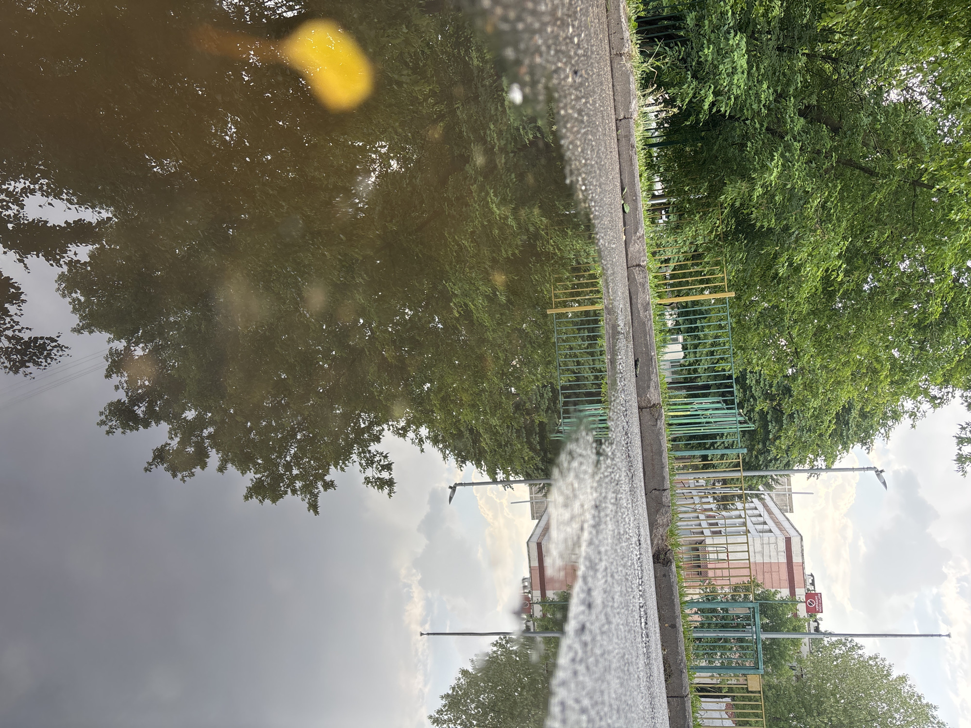 A low-angle, eye-level shot captures a puddle on an asphalt street in Moscow, Russia, reflecting the surrounding environment. The puddle occupies the foreground and extends into the mid-ground, its surface disturbed by ripples, creating a distorted reflection of trees, a colorful metal fence, and a building. The fence, with its posts painted green, yellow, and red, runs along the edge of the sidewalk behind the puddle. Beyond the fence, a building with a white and pink facade stands tall, with windows visible. Two lampposts are present, one with a bird perched on it, and a sign with red lettering is mounted on the other. Lush green trees line the street, their leaves contributing to the dense canopy visible in the reflection. The sky above is partly cloudy, with patches of blue peeking through the white clouds, also reflected in the water. A single, bright yellow leaf floats on the surface of the puddle, adding a touch of color to the scene. The street itself appears wet and textured, with a cracked concrete curb separating the asphalt from a patch of green grass.