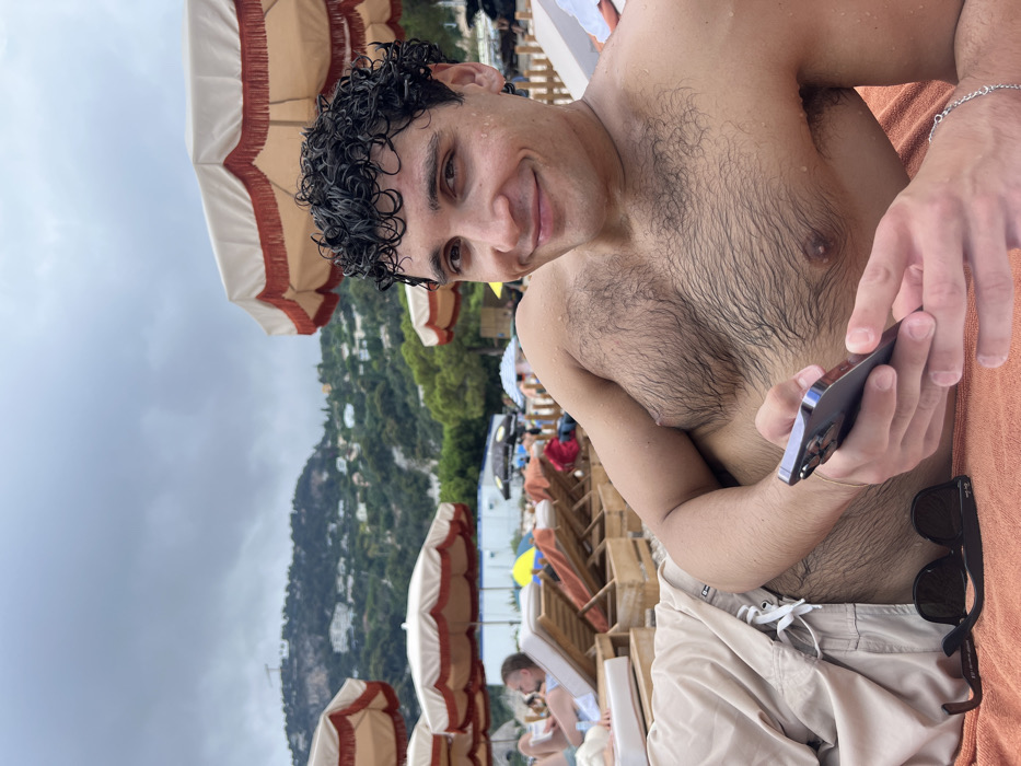 A man with curly wet hair smiles at the camera while sitting on a beach lounge chair. He is shirtless, wearing light-colored shorts, and holding a phone. A pair of sunglasses rests nearby on an orange towel. Beach umbrellas dot the scene, and a green, mountainous landscape is visible in the background under a cloudy sky. Other beachgoers are seated on lounge chairs in the background.