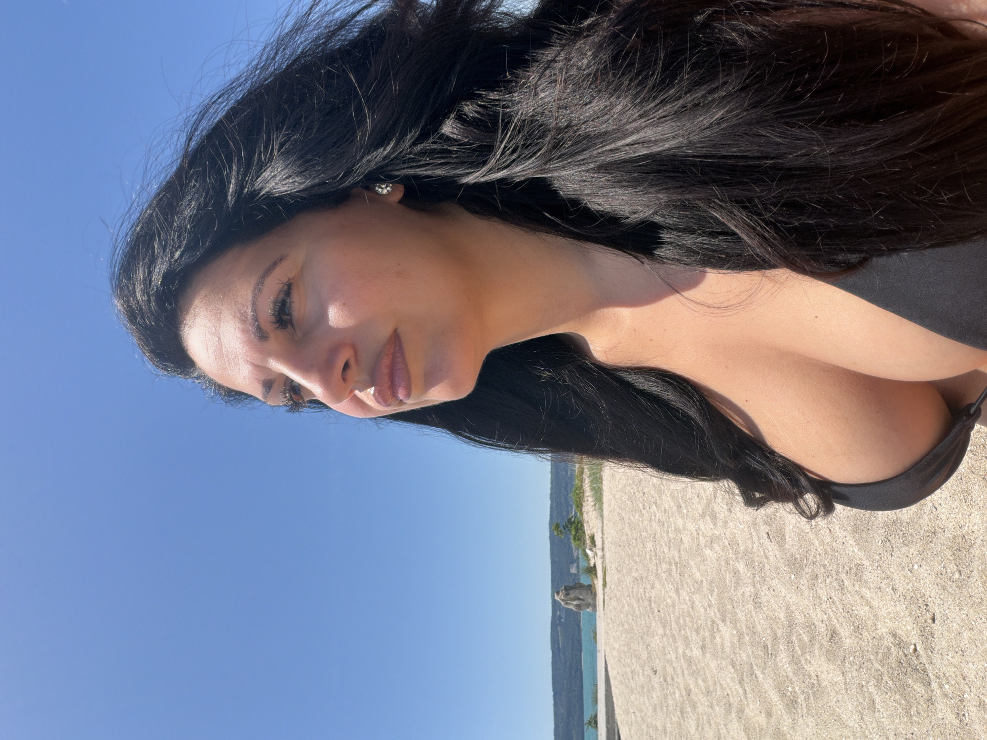 A woman with long, dark hair is pictured outdoors on a sunny day. She is wearing a dark-colored bikini top. Her face is turned to the side, showing her profile. Her hair is blowing in the wind, framing her face. She is wearing small earrings. The background consists of a sandy beach and a body of water, likely the sea, under a clear blue sky. There appears to be a rocky outcrop in the distance on the coastline. The image seems to be taken from a low angle, looking up at the woman. The lighting suggests it is daytime, possibly late morning or early afternoon, with the sun casting shadows and highlighting her skin. The overall mood is relaxed and casual, indicative of a beach setting. No text is visible in the image. The location context is Varna, Bulgaria.
