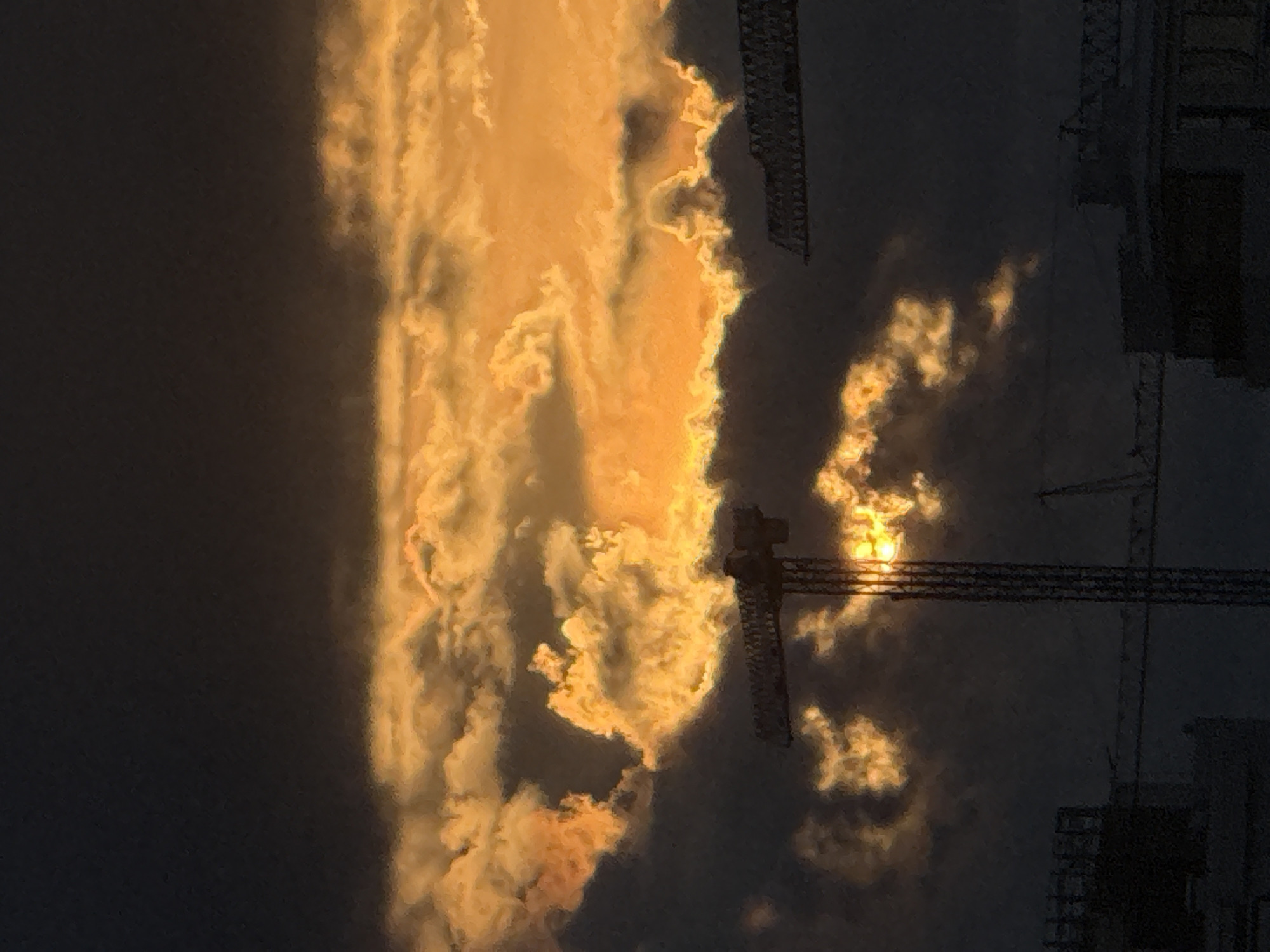 This media file captures a dramatic sunset over Sofia, Bulgaria, as viewed from a construction site. The sky is ablaze with vibrant orange and yellow clouds, which are illuminated by the setting sun. The sun itself is visible as a bright, almost ethereal orb peeking through the textured clouds.
The scene is dominated by the silhouettes of construction cranes and partially built structures, characteristic of an urban landscape undergoing development. These man-made objects contrast sharply with the natural beauty of the sunset, creating a visually striking juxtaposition. The dark, almost black sky serves as a dramatic backdrop, accentuating the intense colors of the clouds and the sun.
The time of day is clearly sunset, indicated by the low angle of the sun and the rich, warm hues in the sky. The weather appears clear, allowing for an unobstructed view of the celestial display. The presence of construction cranes strongly suggests an urban environment, and the provided location context confirms this is Sofia, Bulgaria.
No people are visible in the image. The mood evoked by the image is one of awe and wonder, with the powerful natural phenomenon of the sunset overwhelming the industrial elements of the scene. There is no visible text in the media file.